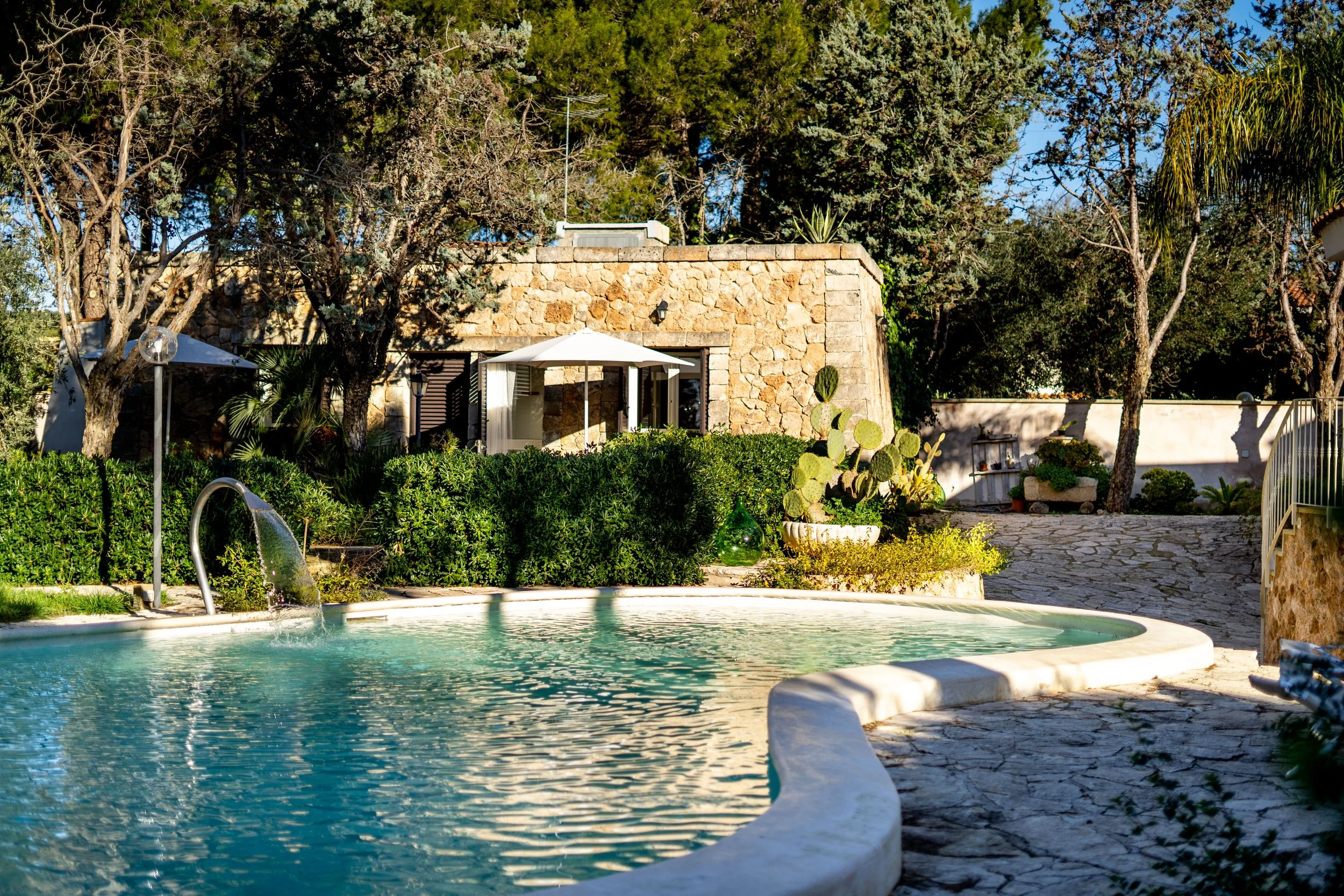 Ein Swimmingpool mit umgebenden Pflanzen und einer Steinhütte im Hintergrund, sonniger Gartenbereich