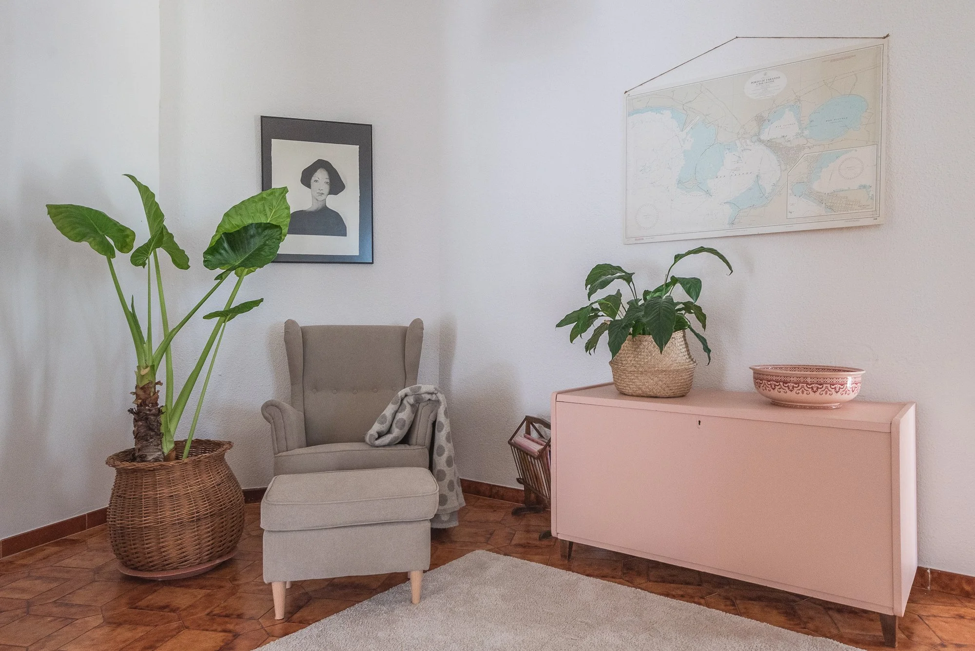Ein Wohnzimmer mit braunem Holzboden, einer großen Zimmerpflanze in einem Korb, einem Beistuhl mit Decke, einer pinkfarbenen Kommode, zwei Wandbilder, einem größeren mit einer Landkarte und einem kleineren mit einem schwarzen und weißen Porträt einer