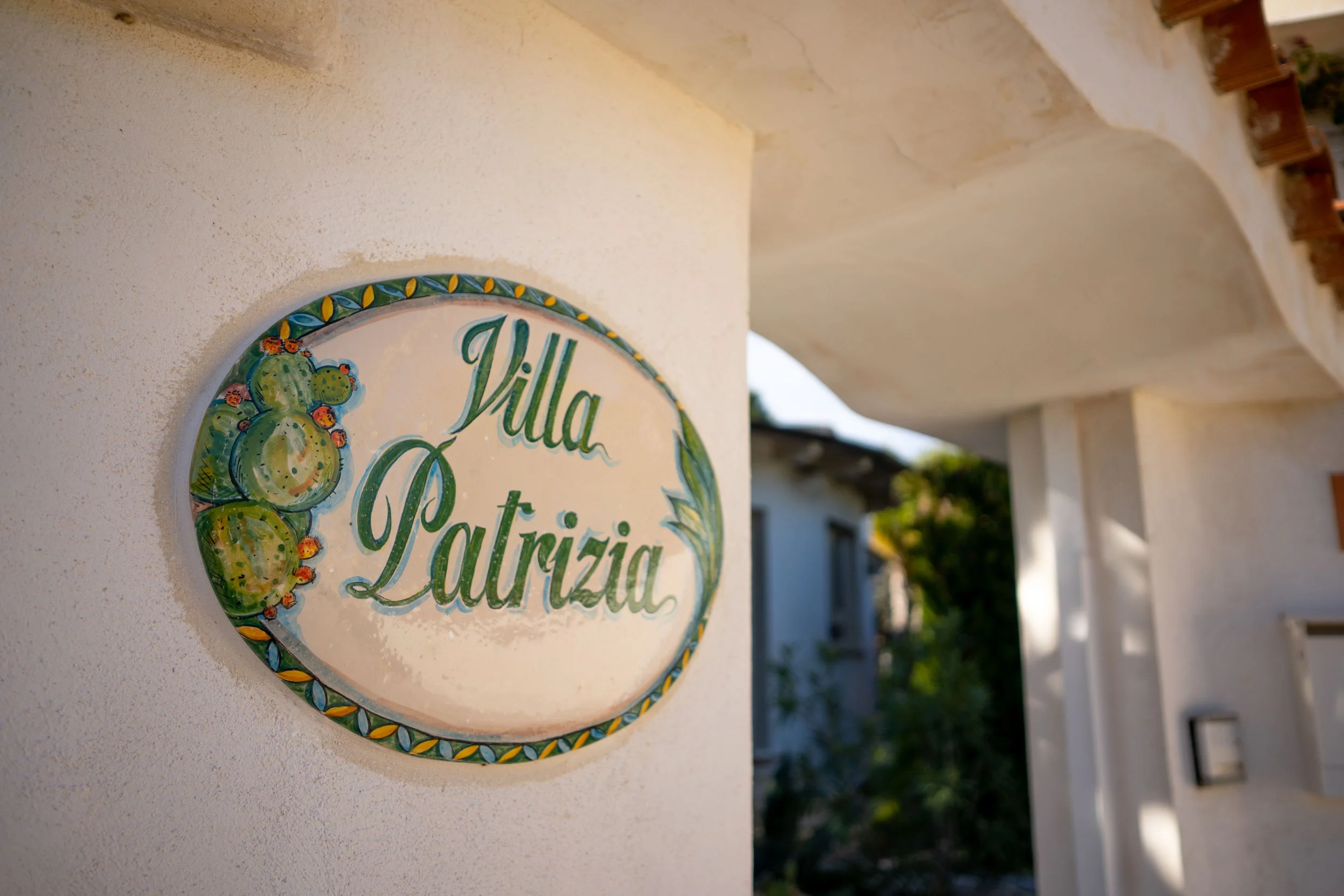 Blaue und grüne beschilderte Tafel mit der Aufschrift Villa Patrizia, umgeben von einem dekorativen Rahmen mit Kaktus-Illustration, an einer weißen Wand befestigt.