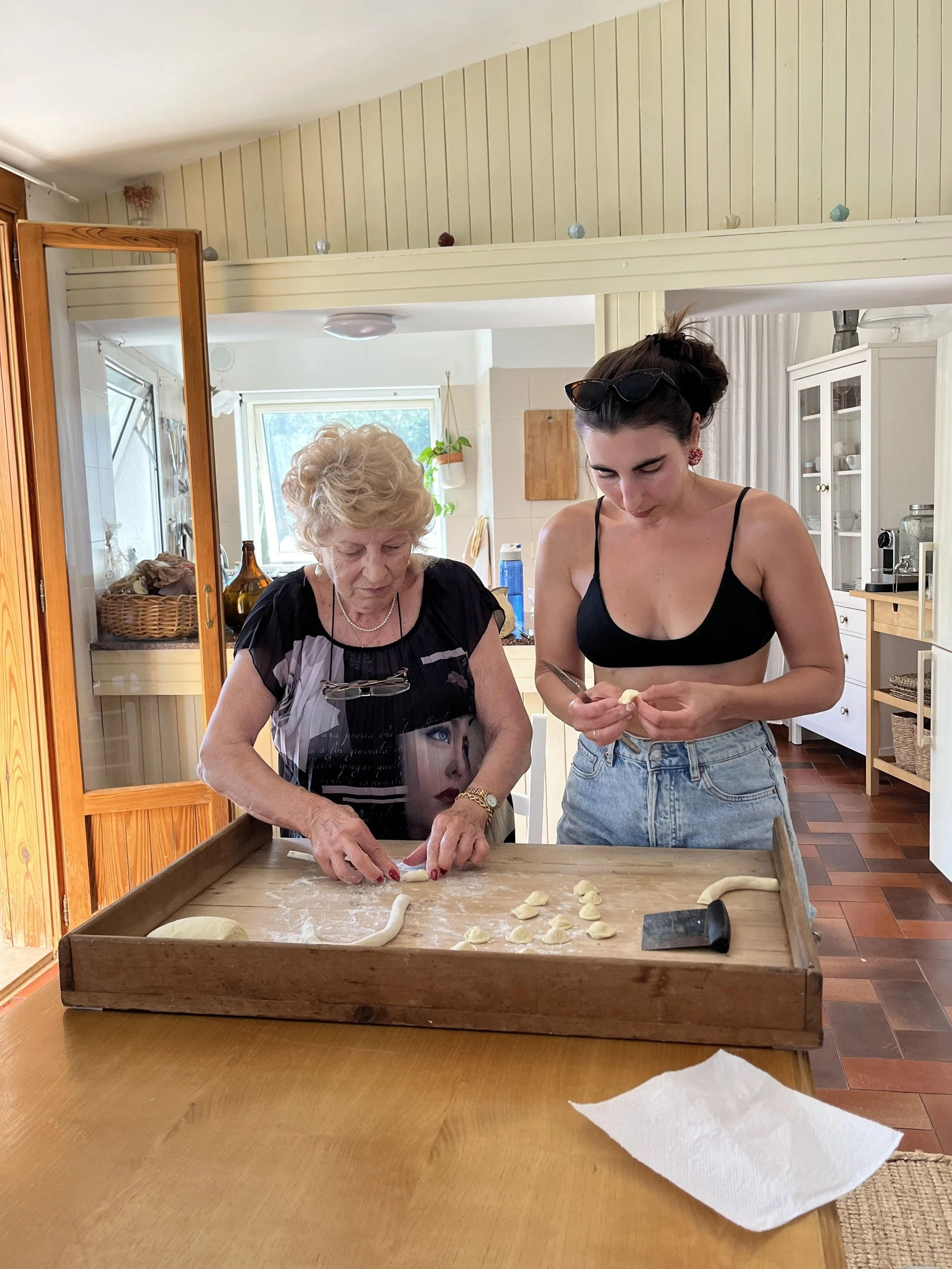 Zwei Frauen beim Teigkneten in einer Küche, eine ältere Frau und eine jüngere Frau, im Vordergrund ein Tisch mit Teigresten und einem Messer.