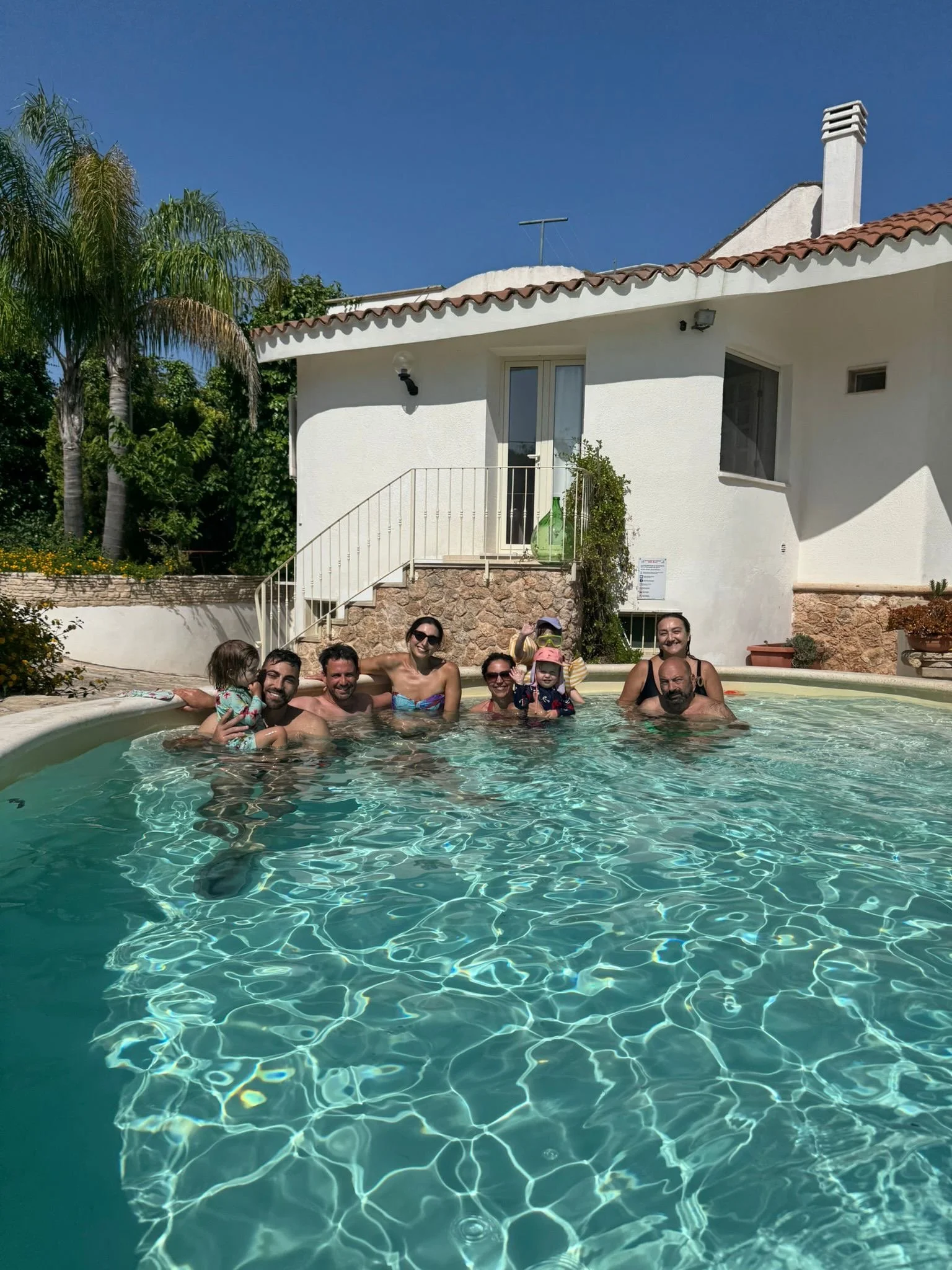 Eine Gruppe von Erwachsenen und Kindern schwimmen und sitzen im Pool vor einem weißen Haus mit roten Ziegeldach, umgeben von Pflanzen und Palmen, bei sonnigem Wetter.