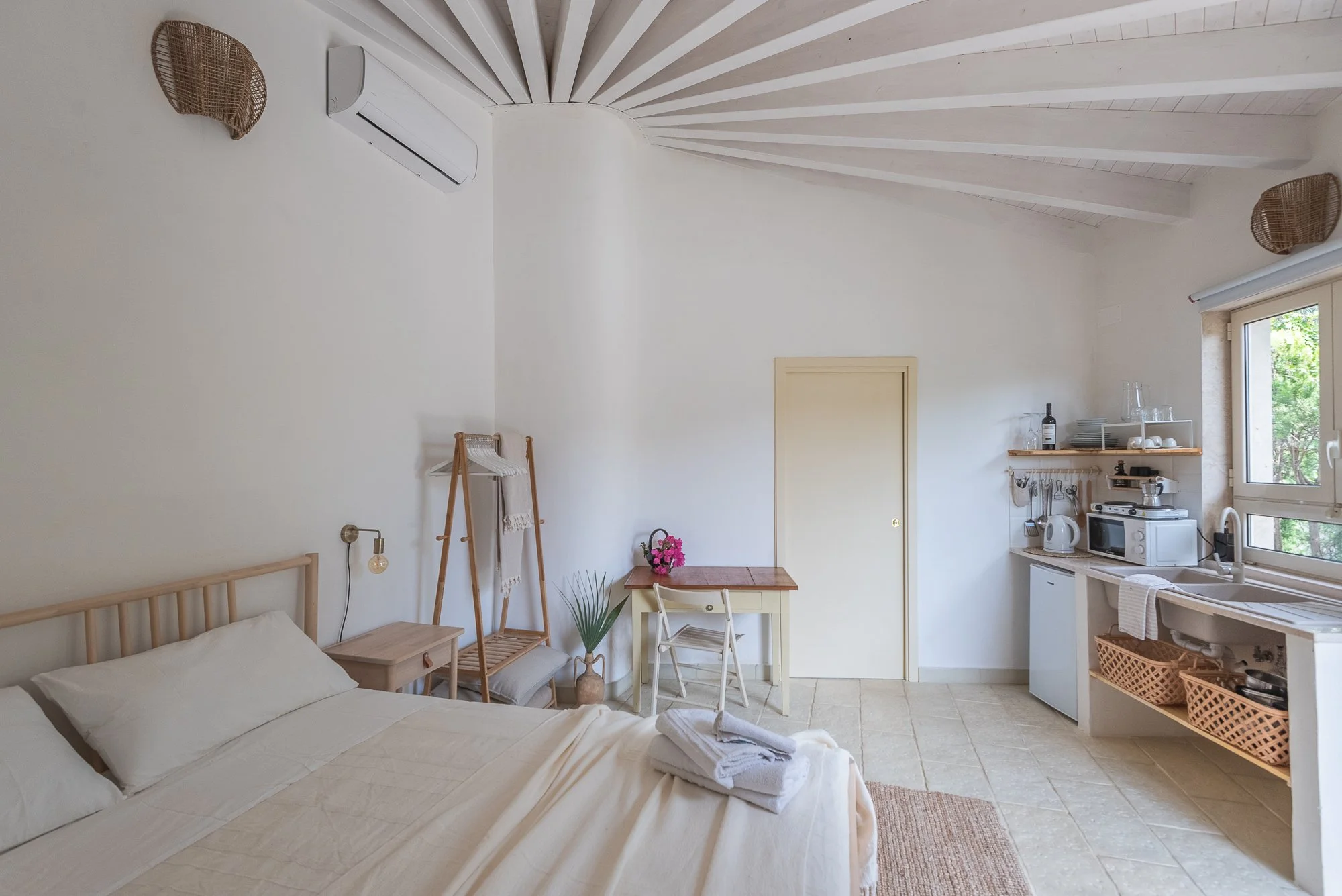 Ein minimalistisches, helles Schlafzimmer mit offener Küche, weißem Bett, Fenster mit Blick auf grüne Bäume, Holzdecke mit Dachbalken und ländlichem Dekor.