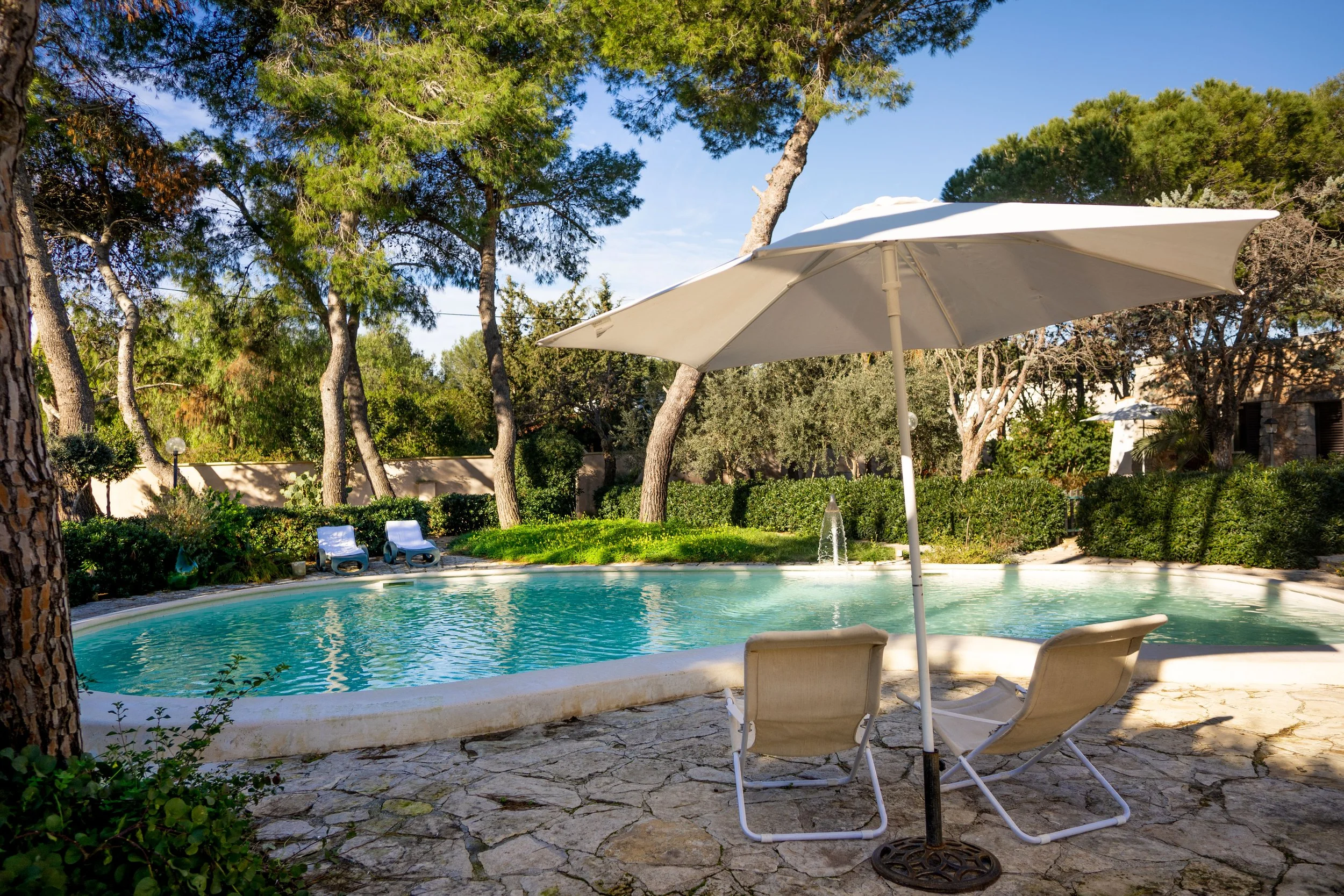 Ein Swimmingpool im Freien, umgeben von Bäumen, mit zwei Liegestühlen und einem Sonnenschirm im Vordergrund, und zwei weitere Liegestühle am Rand des Pools im Hintergrund.