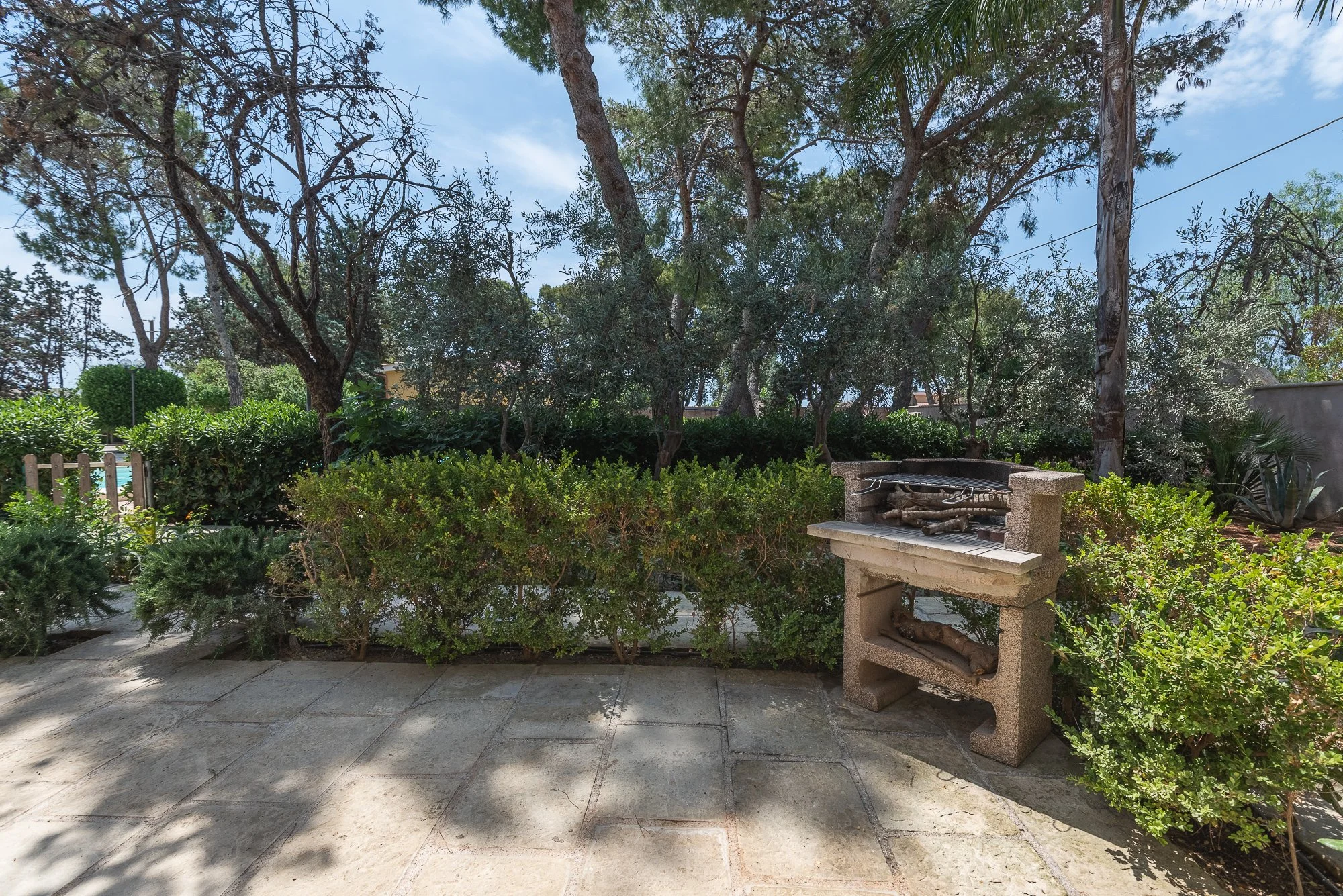 Garten mit Grill auf gepflastertem Boden, umgeben von grünen Sträuchern und Bäumen, blauer Himmel.