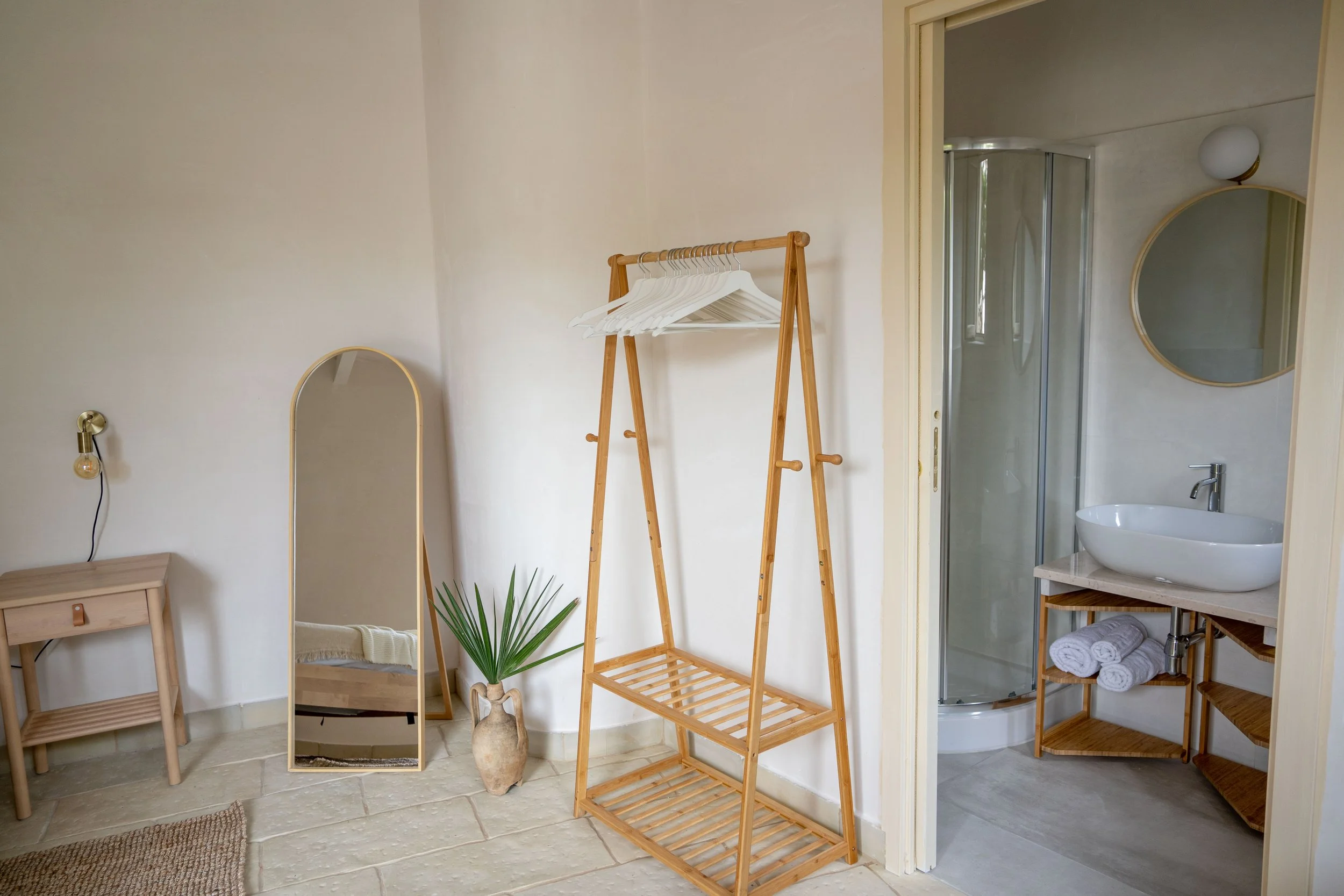 Ein heller Raum mit Wandspiegel, Holztablett, Pflanzen, Kleiderständer mit Hängekleidern, und einem Badezimmer mit Waschbecken und Dusche.