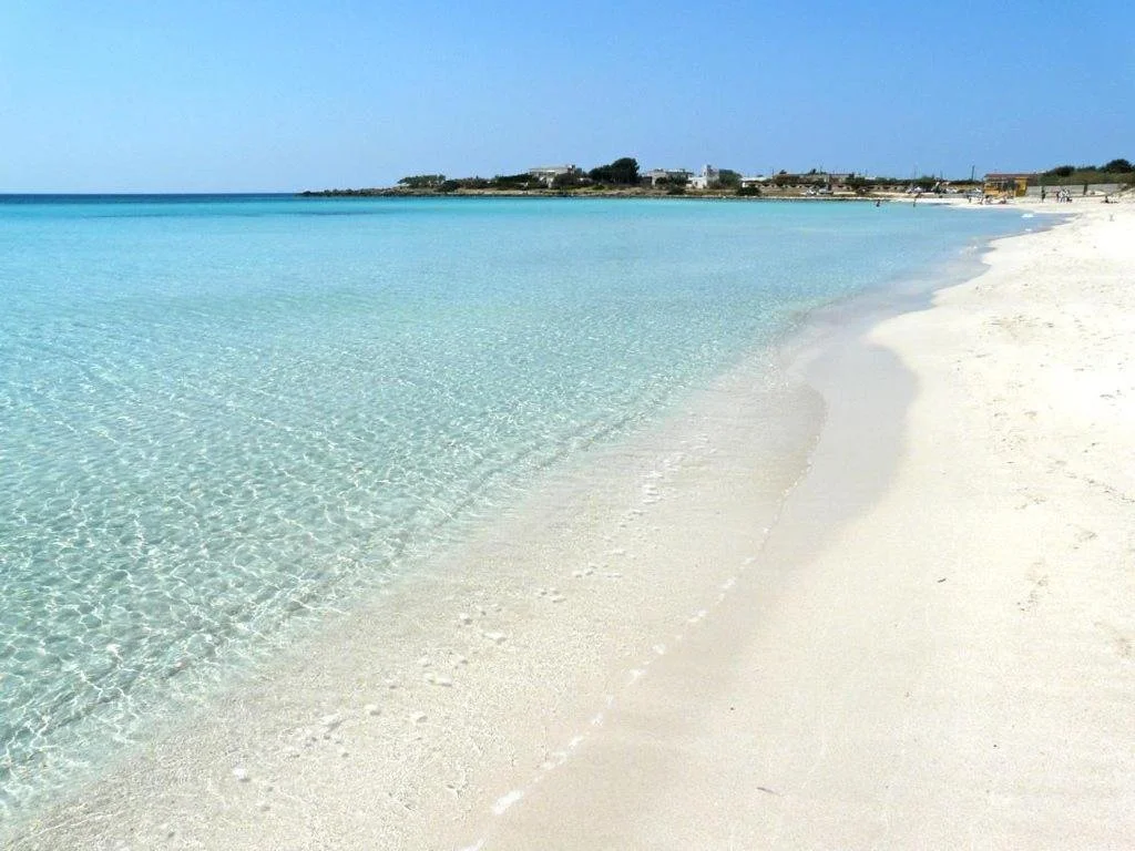 Strand mit weißem Sand und klarem türkisfarbenem Wasser, im Hintergrund einige Gebäude und Bäume, blauer Himmel