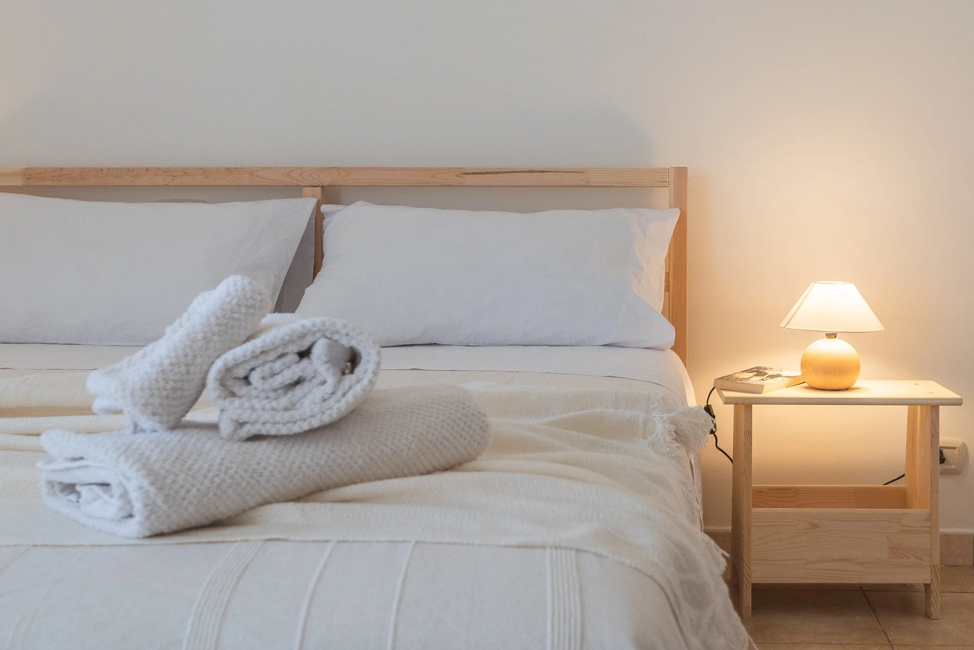 Bett mit weißen Handtüchern und Kissen, Nachttisch mit Lampe und Buch, gemütliches Schlafzimmer mit warmem Licht.