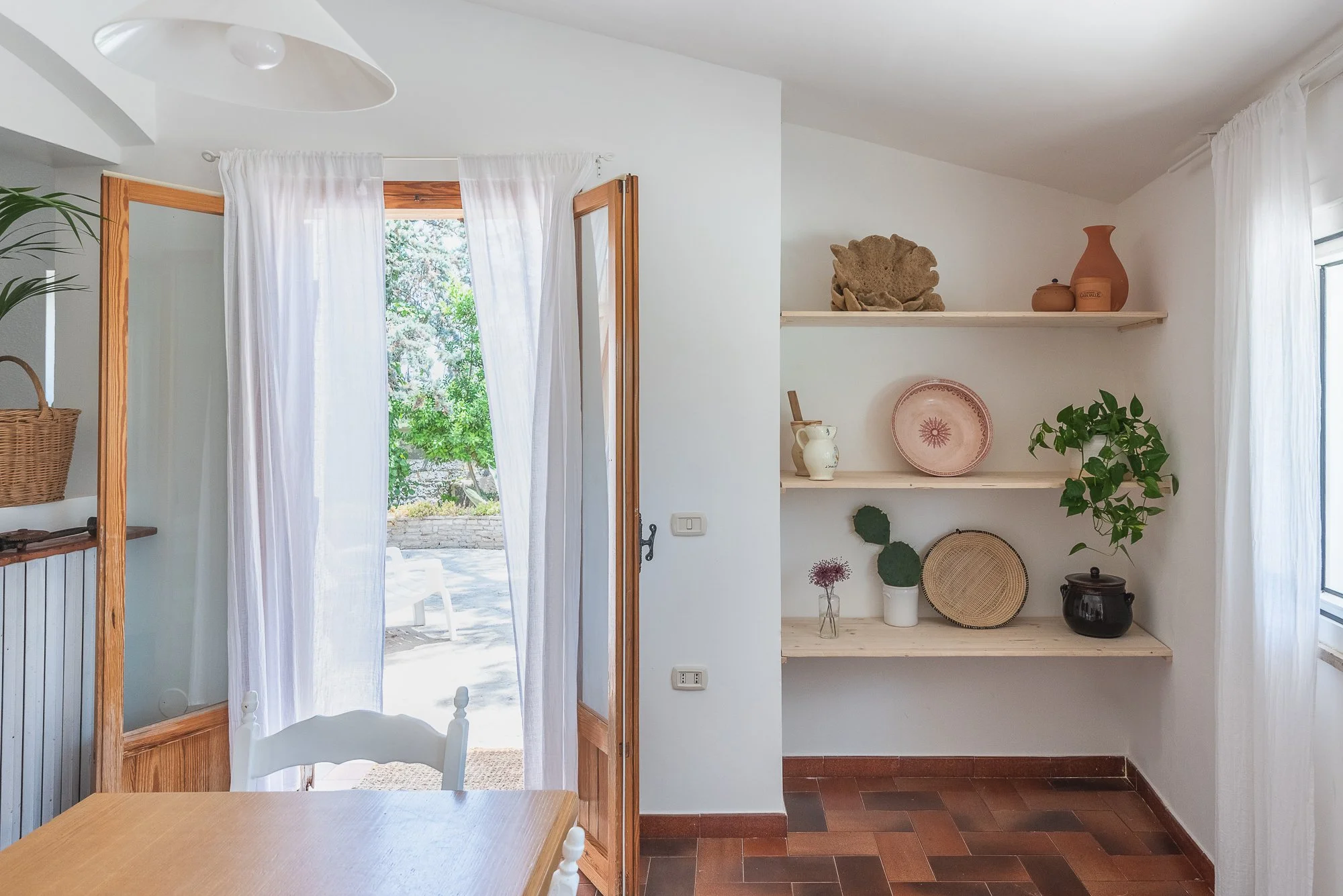 Ein heller Raum mit offener Tür, der nach draußen führt, und Holzregalen, die mit dekorativen Keramiken, Pflanzen und Körben bestückt sind.