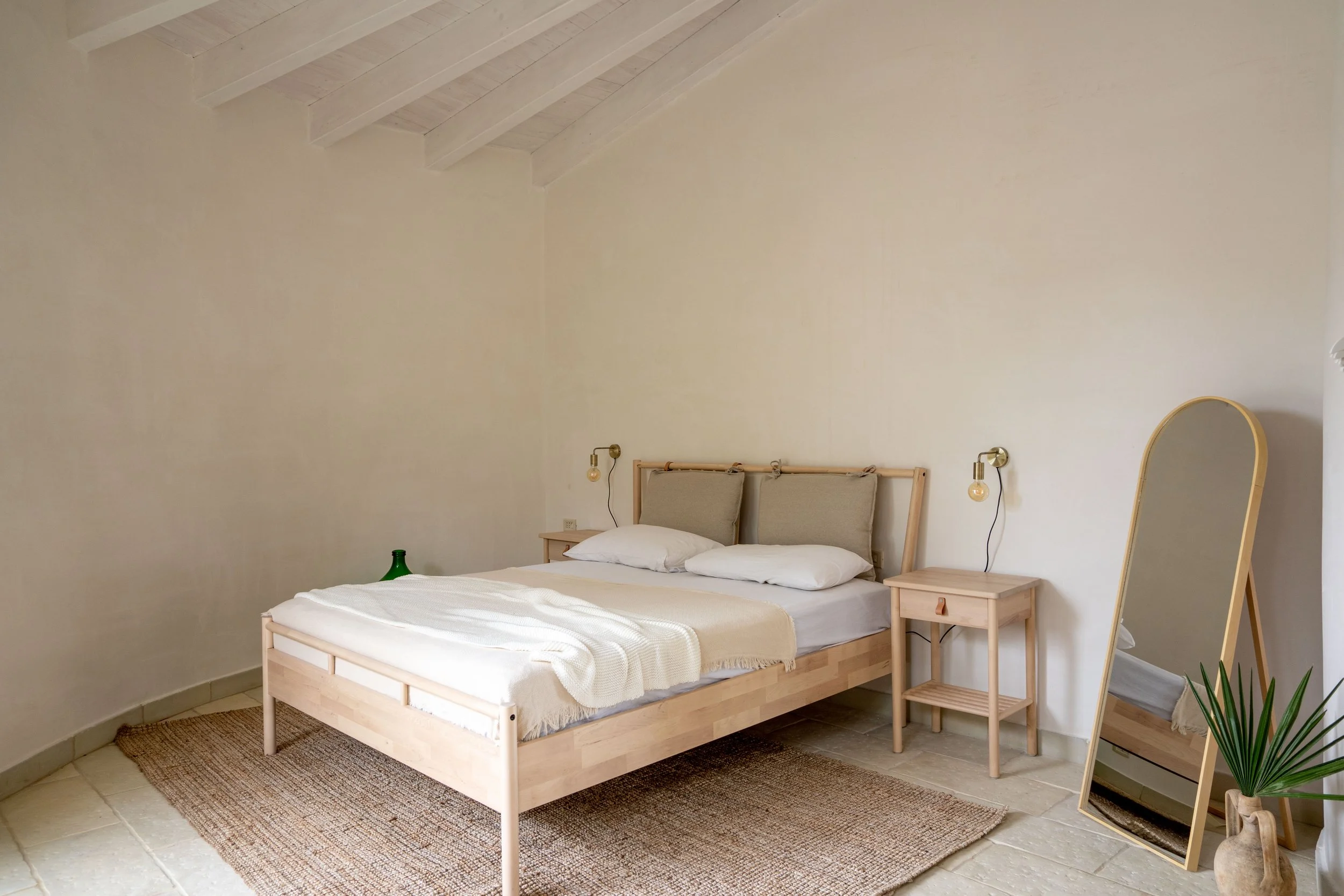 Schlafzimmer mit einfachem Holzbett, zwei Nachttischen, wandhängenden Lampen, großen Spiegel und Pflanzen in einem minimalistischen, hellen Raum.