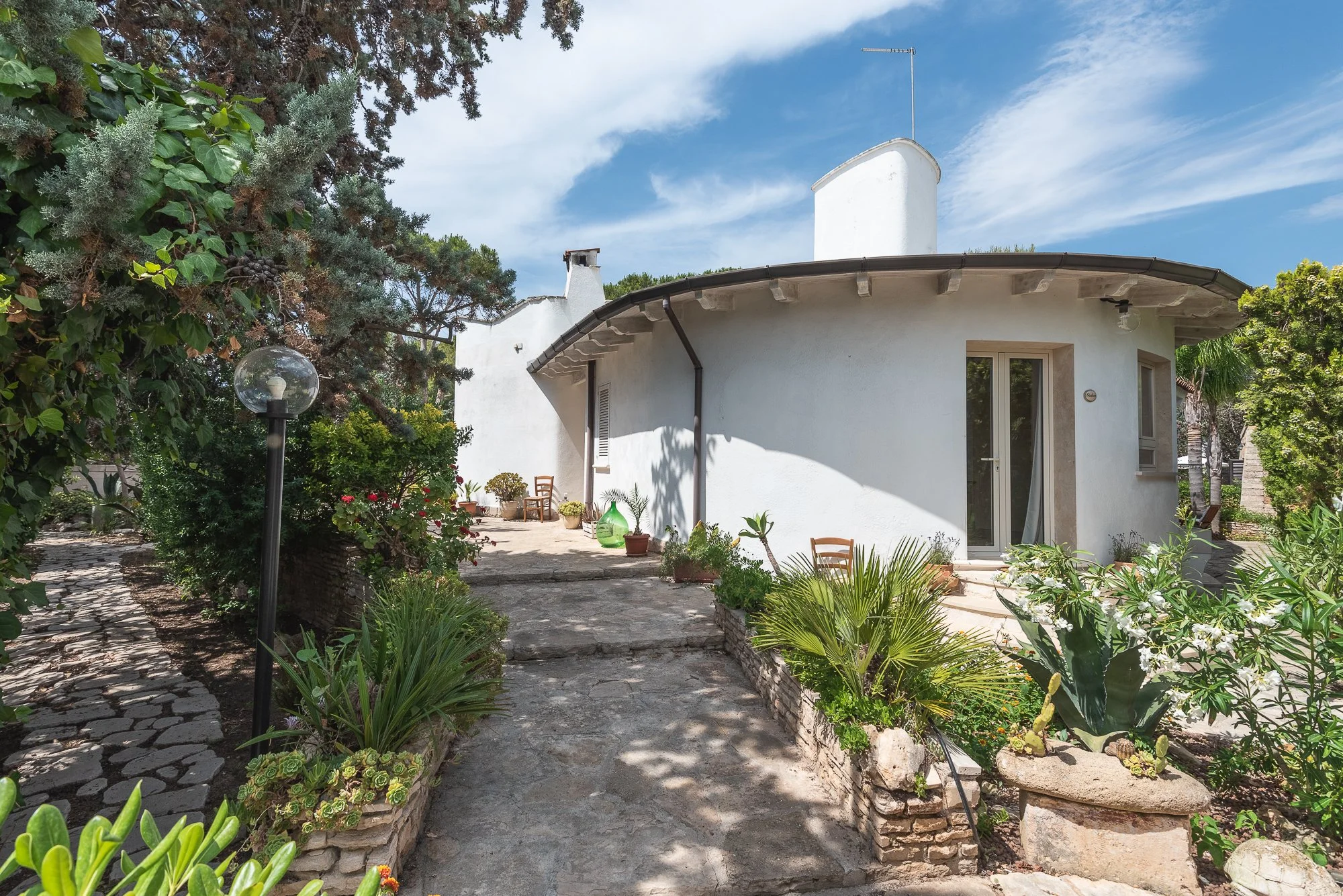 Weiße, runde Haus mit Terrassentür und Pflanzen im Vorgarten unter blauem Himmel.
