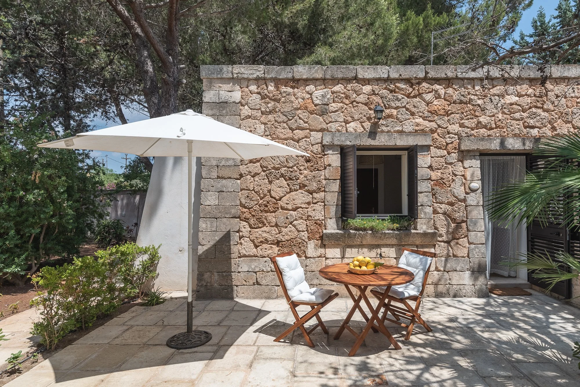 Gartenbereich mit Steinhaus, Sonnenschirm, Rattanstisch, Stühlen und Obstschale, im Freien bei Sonnenschein.