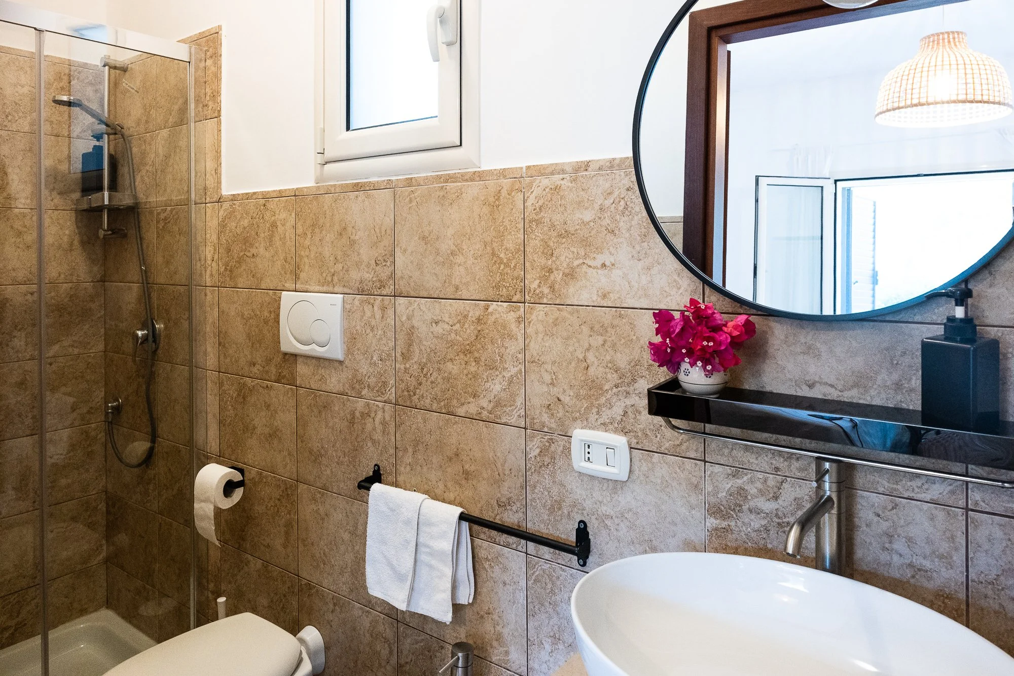 Badezimmer mit beigen Fliesen, Dusche, rundem Spiegel, Fenster, Waschbecken, Handtuchhalter mit Handtuch, Blumentopf mit pinken Blumen, Seifenspender.