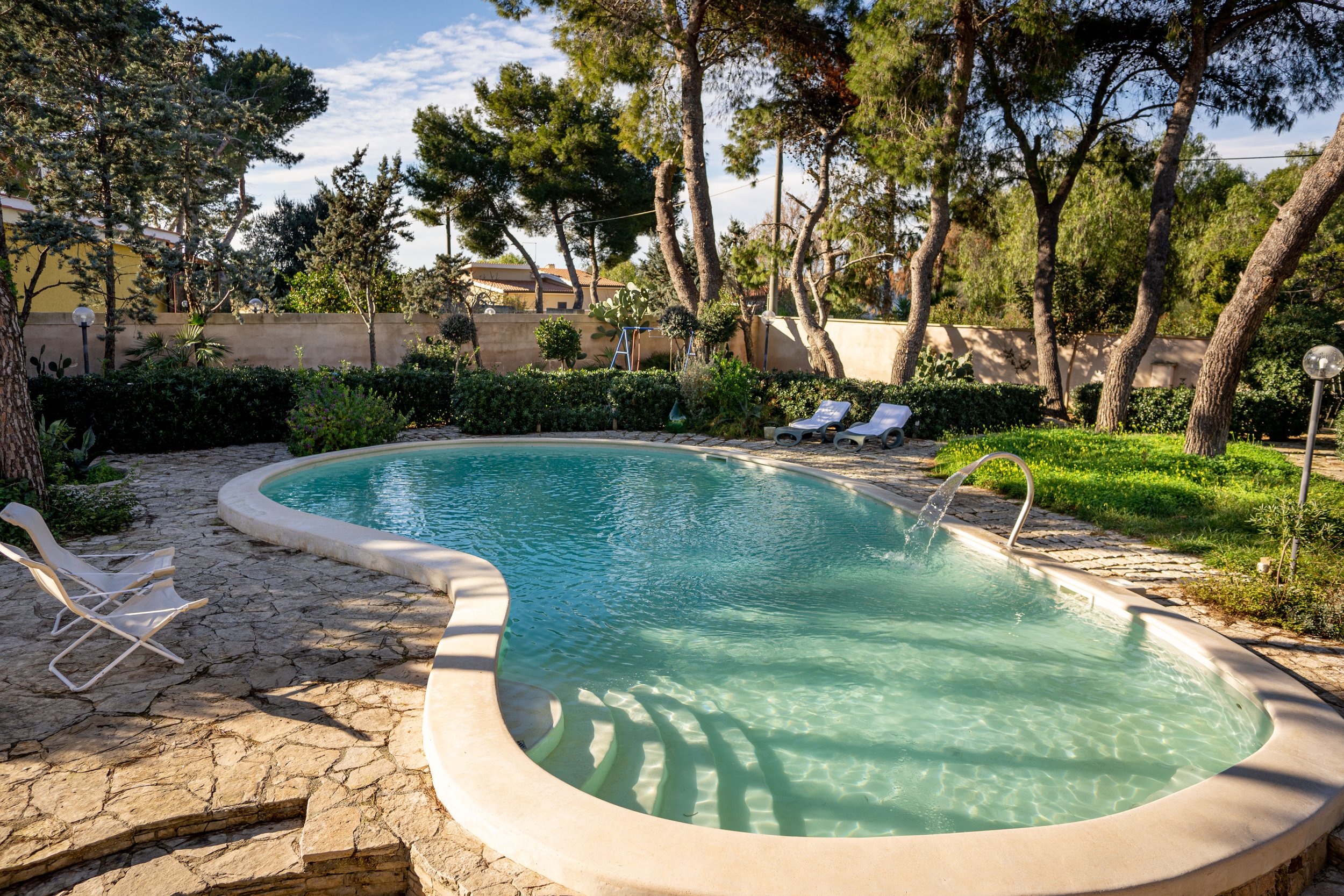 Ein freundlicher Pool in einem Garten mit Bänken, umgeben von Bäumen and Sträuchern, bei sonnigem Wetter.