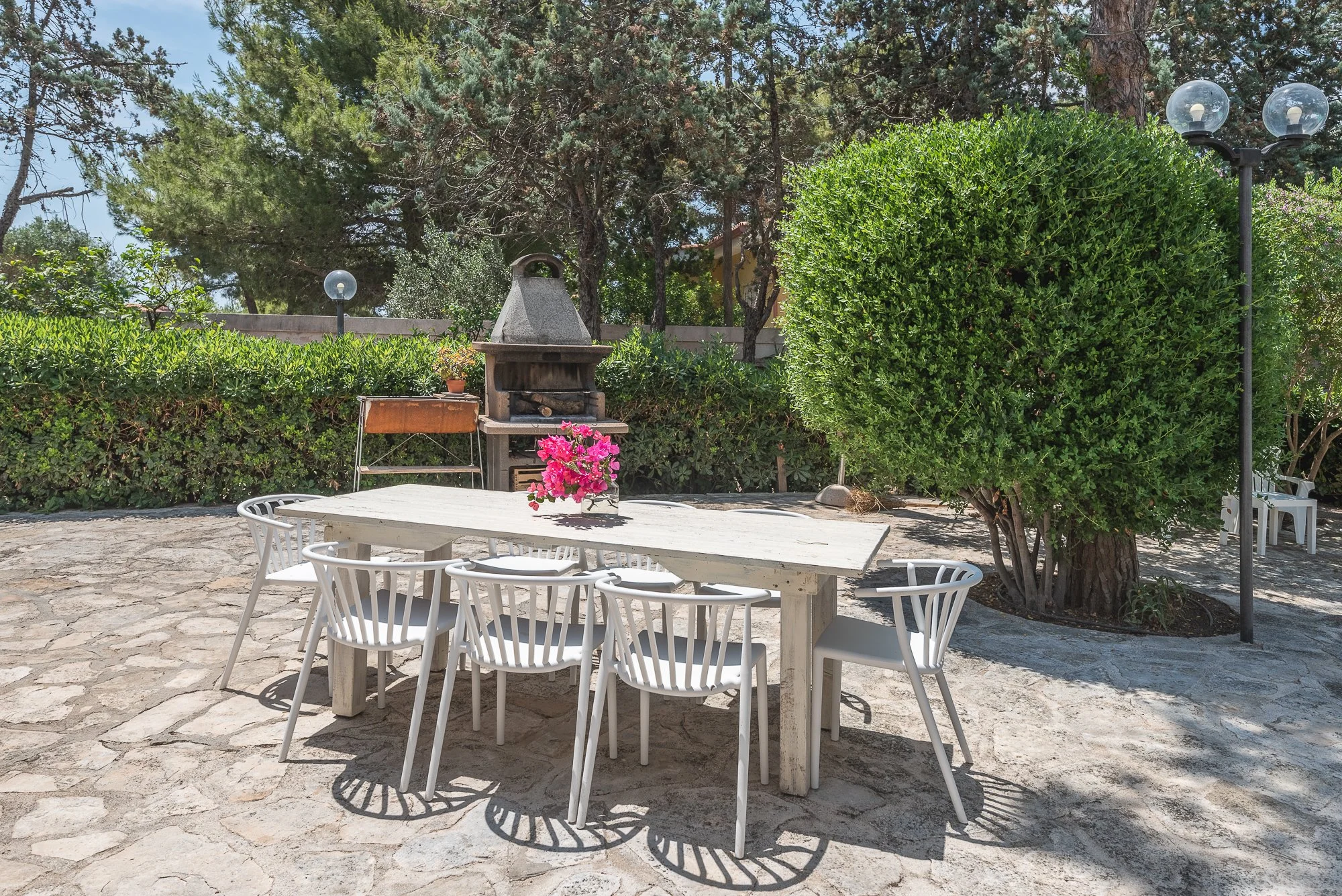 Ein weißer Esstisch mit sechs weißen Stühlen im Garten mit großer Baum, Sträuchern und einem Grill in der Hintergrund, Sonnenschein und Schatten. Auf dem Tisch eine Vase mit pinken Blumen.