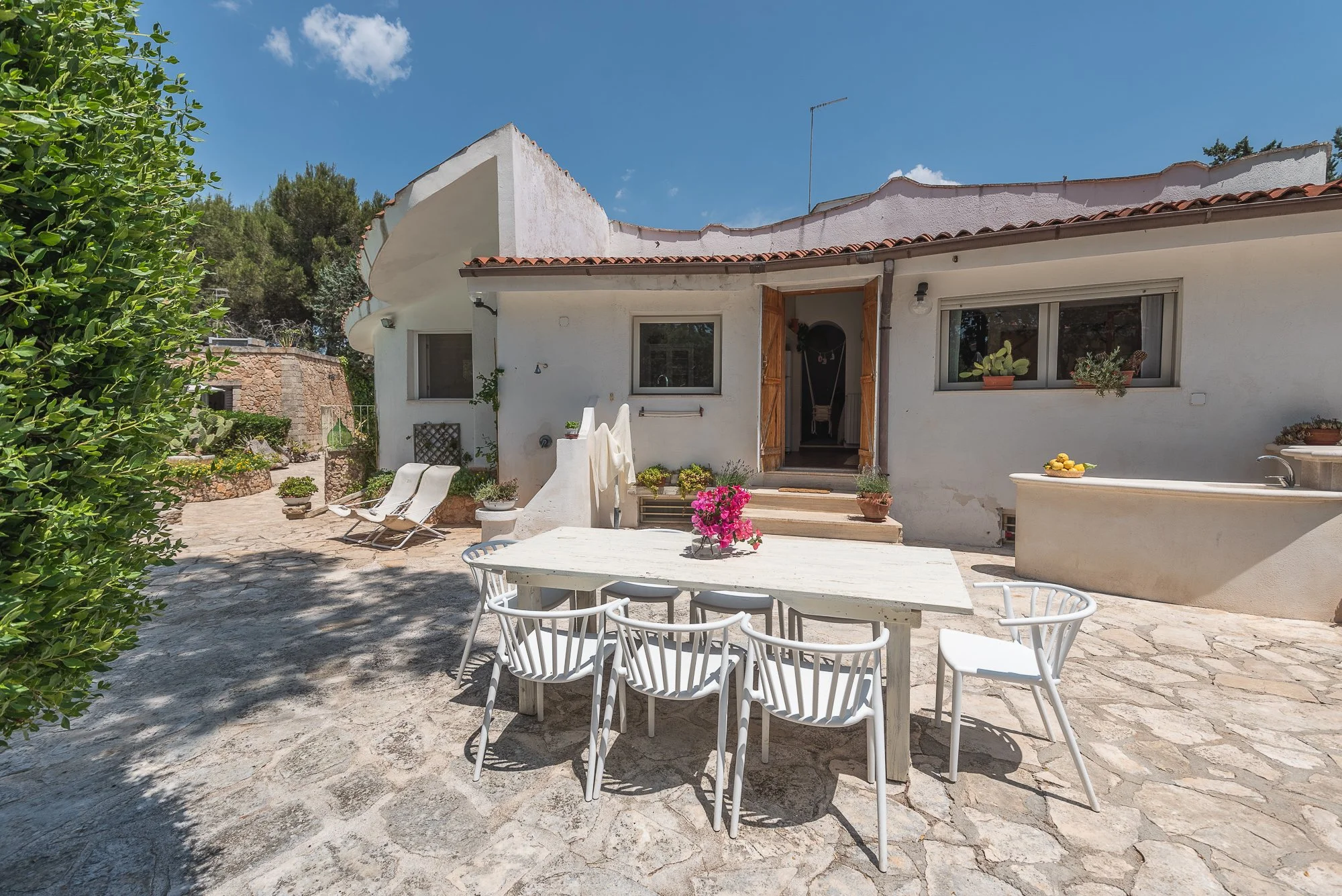 Ein heller Außenbereich eines weißen mediterranen Hauses mit Terrassenmöbeln, Pflanzen und einem blauen Himmel.