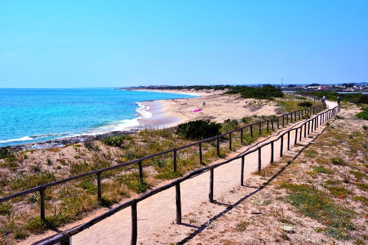 Ein Strand mit Sand, Wasser und einer promenade mit Holzgeländer. Es gibt wenige Menschen am Strand und eine pinke Strandmatte liegt auf dem Sand. Der Himmel ist klar und blau.