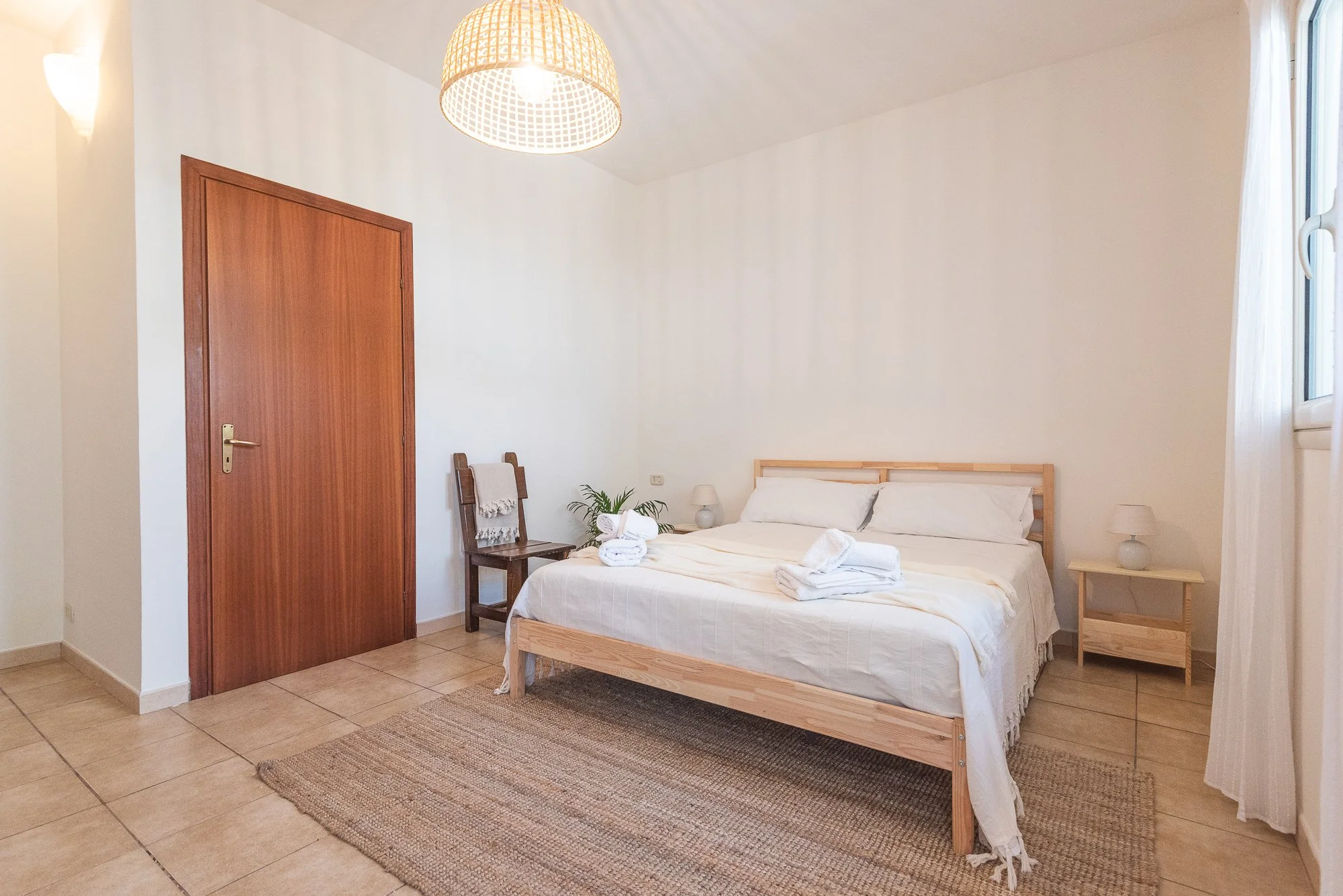 Schlafzimmer mit Holzbett, weißen Bettwäsche, kleinen Nachttischen mit Lampen, einer Tür, einem Stuhl mit Handtuch, einer Pflanze, einem Teppich und einem Fenster mit weißen Vorhängen.