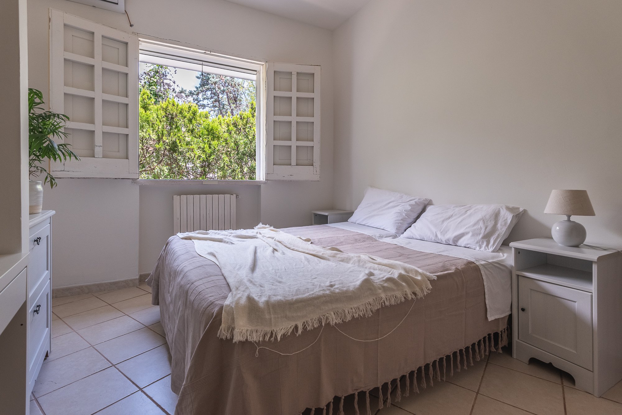 Ein einfach eingerichtetes Schlafzimmer mit einem offenen Fenster, das nach draußen auf Bäume blickt. Ein Bett mit weißen Kissen und Decke, seitliche Nachttische mit Lampe, und eine Pflanze auf einem Schrank.
