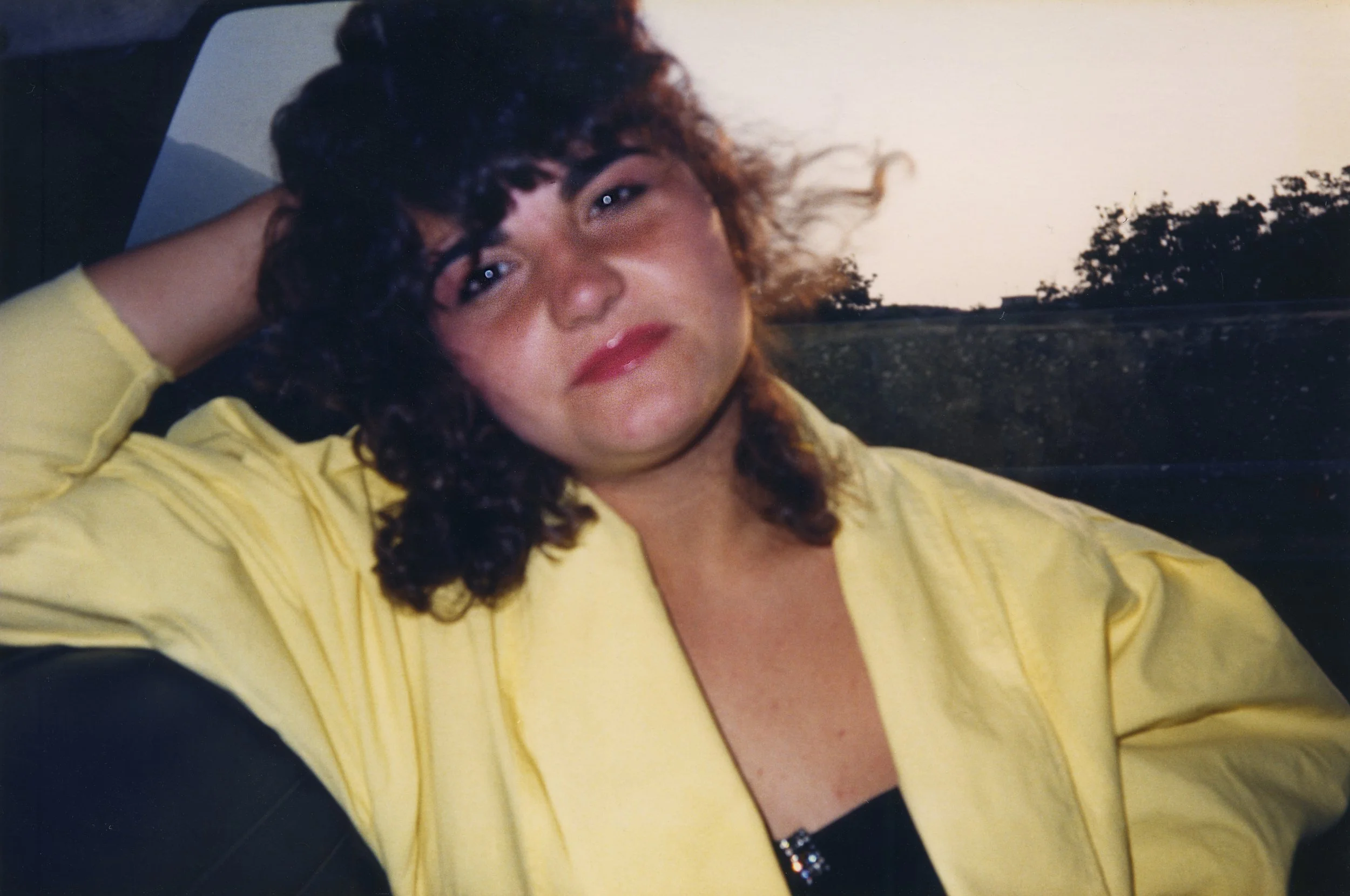 Junge Frau mit lockigem Haar, in gelber Jacke, sitzt im Auto, schaut in die Kamera, bei Sonnenuntergang im Hintergrund.