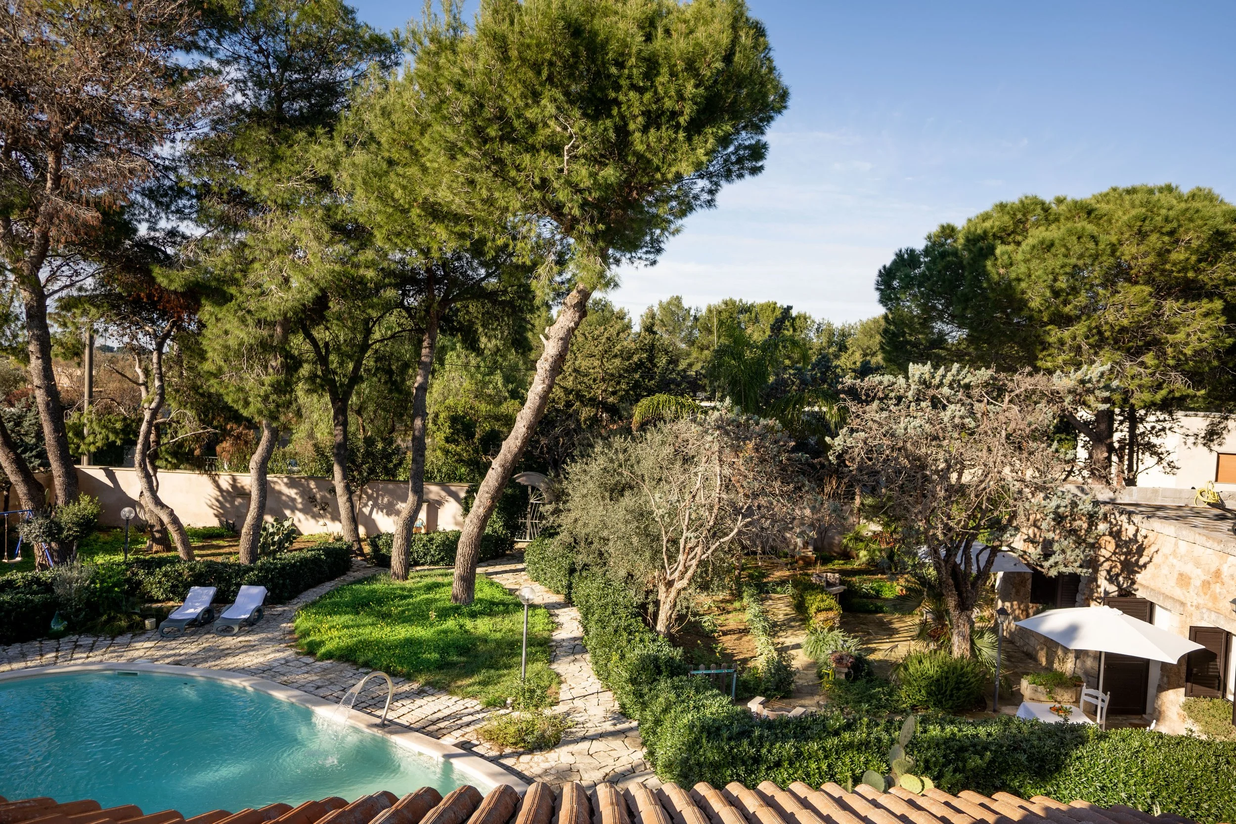 Garten mit Swimmingpool, Liegestühlen, Bäumen, Büschen und einer Terrasse unter einem blauen Himmel.