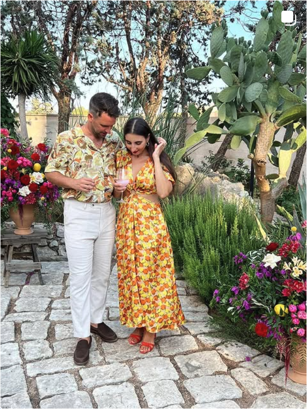 Ein Mann und eine Frau stehen in einem Garten, der mit bunten Blumen und großen Pflanzen geschmückt ist, während sie ein Glas Wein trinken.