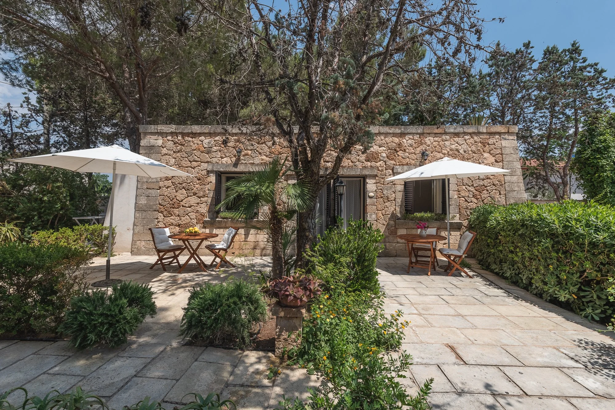 Ein terrassenartiger Garten mit zwei Tischen, weißen Sonnenschirmen, Bänken und einer Steinmauer, umgeben von Büschen und Bäumen, an einem sonnigen Tag.