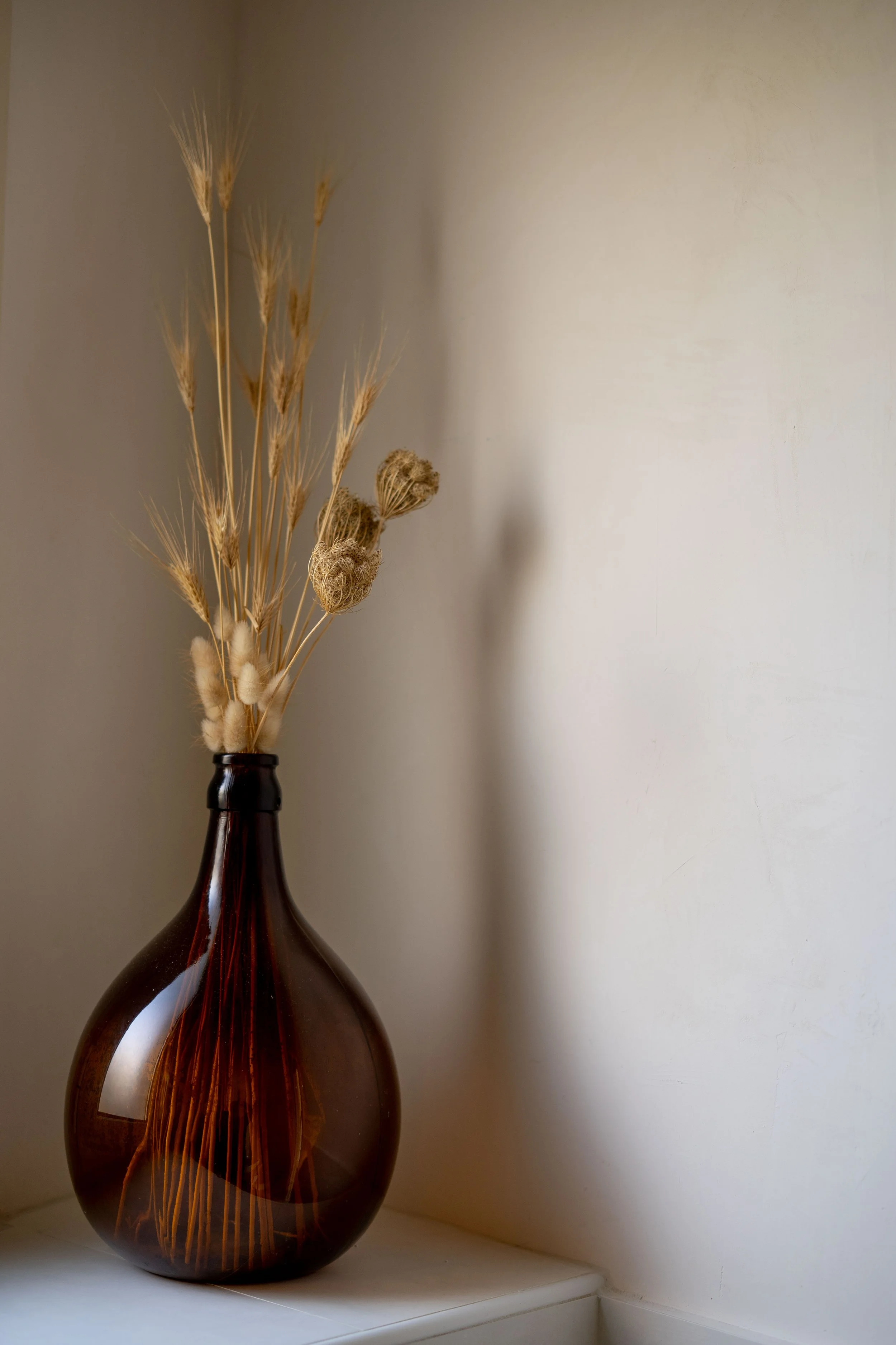 Braune Glasvase mit getrockneten Pflanzen auf einem weißen Tisch an einer hellen Wand