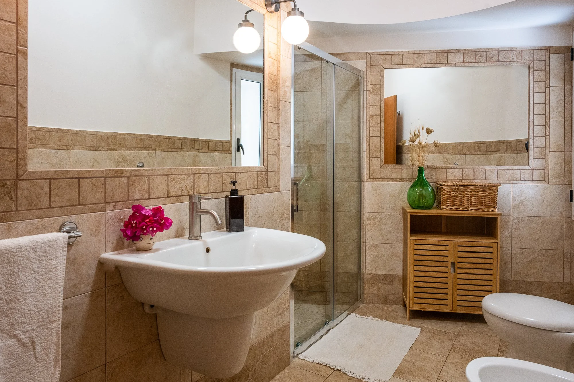 Ein Badezimmer mit einer großen Spiegelwand, Waschbecken, Dusche, Wc, Holzschränkchen, Dekorationsvase, Handtuchhalter mit Handtuch und beige Fliesen.