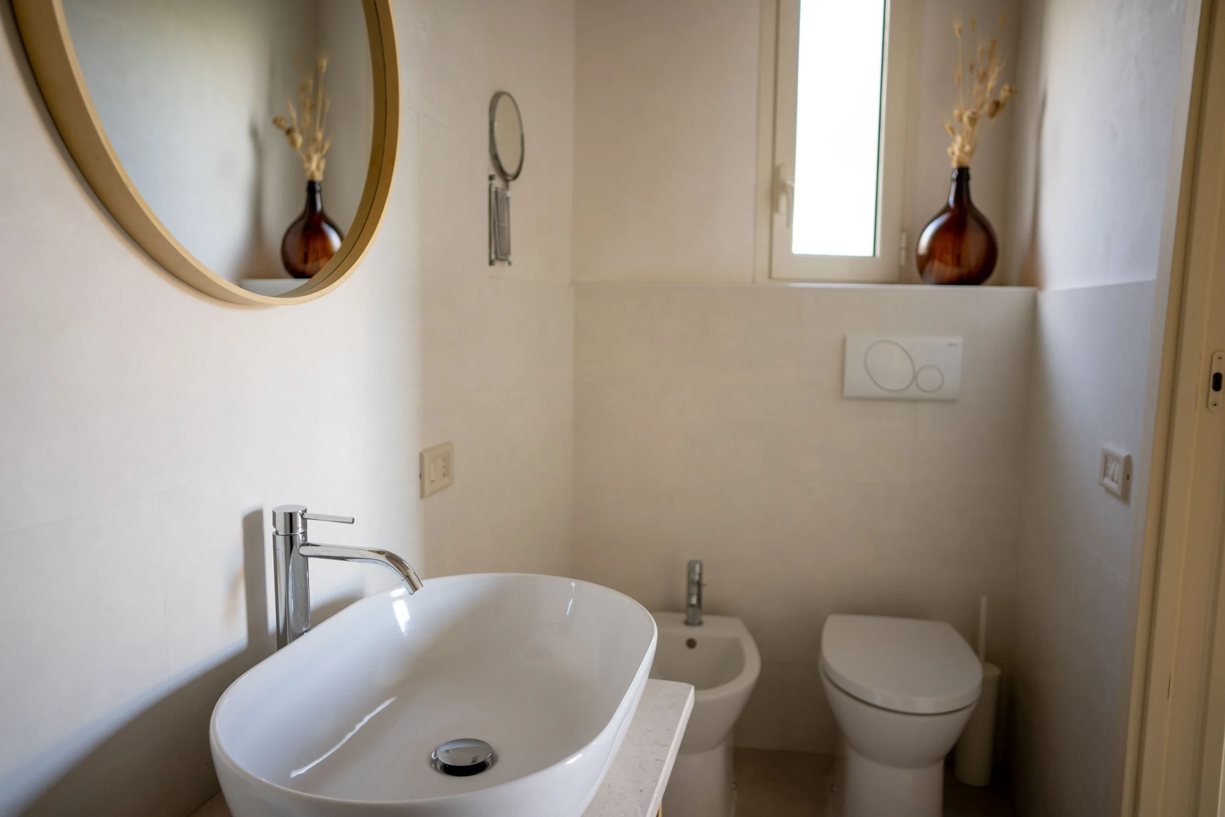 Modernes Badezimmer mit ovalem weißen Waschbecken, Wandspiegel, Bidet, Toilette, Fenster mit Fensterbank mit Vase, helle Wände und minimalistische Dekoration.