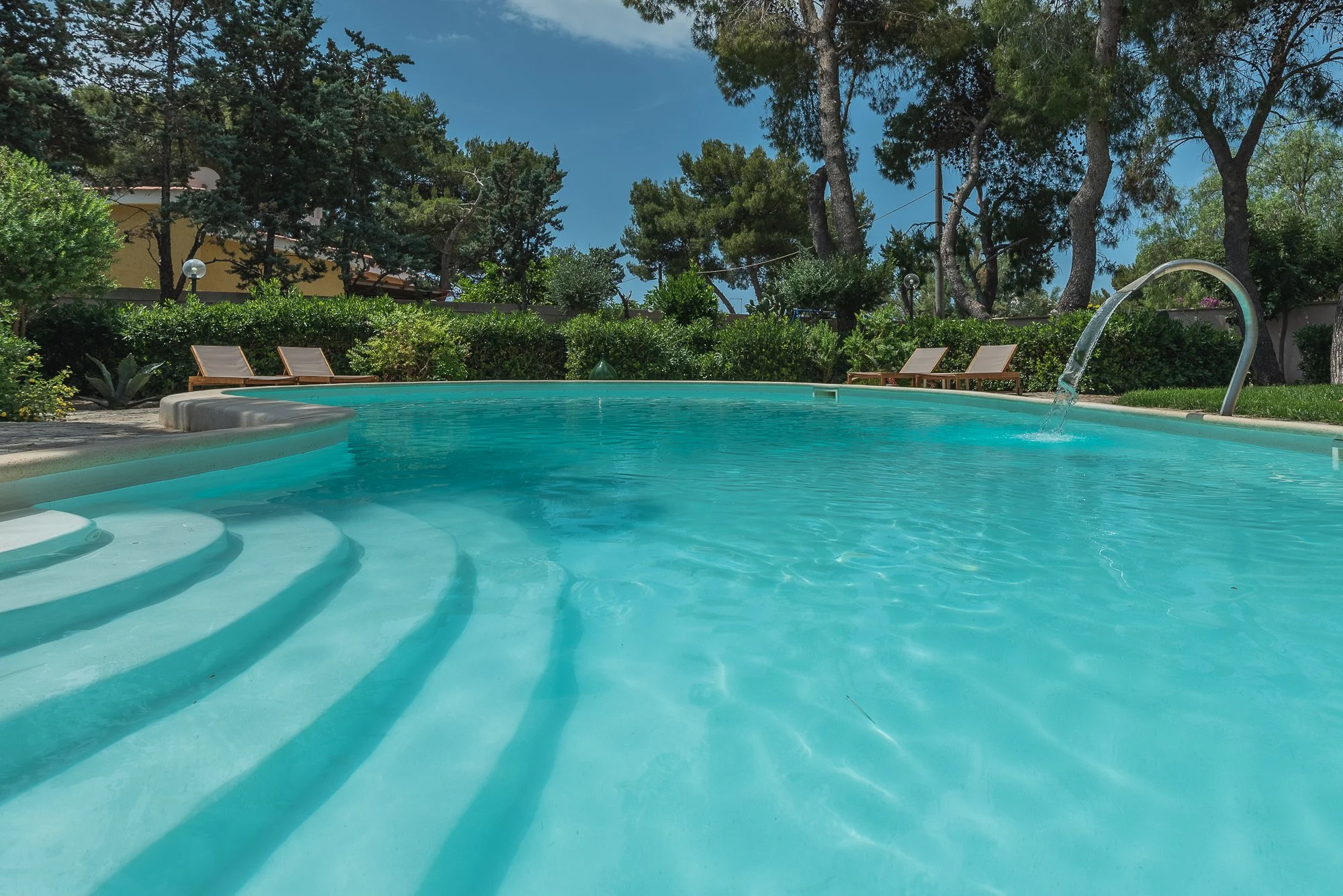 Schwimmbecken im Freien mit Wasserfall, umgeben von Bäumen und Liegestühlen unter blauem Himmel.
