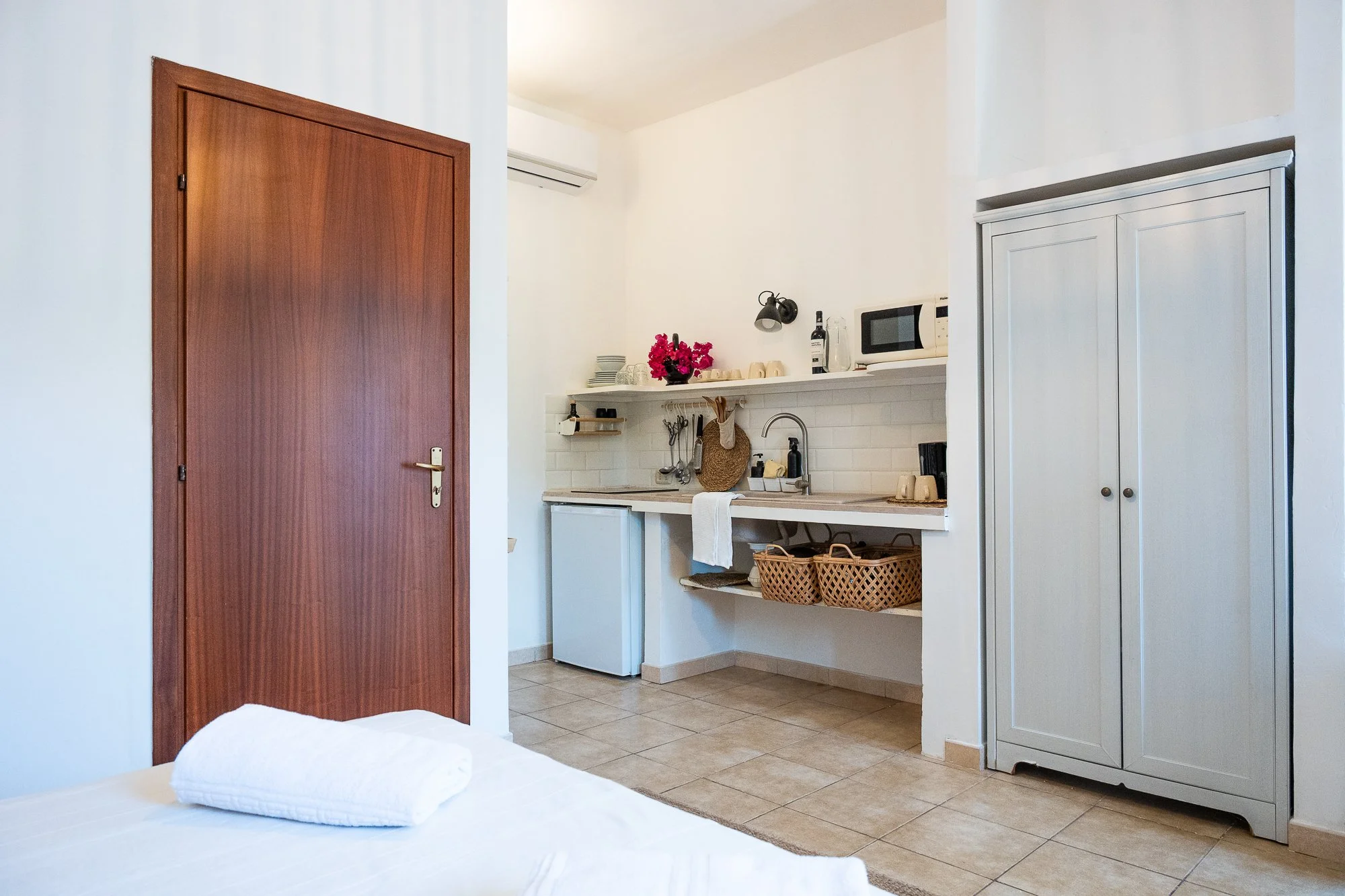 Ein Zimmer mit einem Bett, einer HolzTür, einem weißen Schrank, einer Küchenzeile mit Geschirr und Geräten, sowie ein Handtuch im Vordergrund.