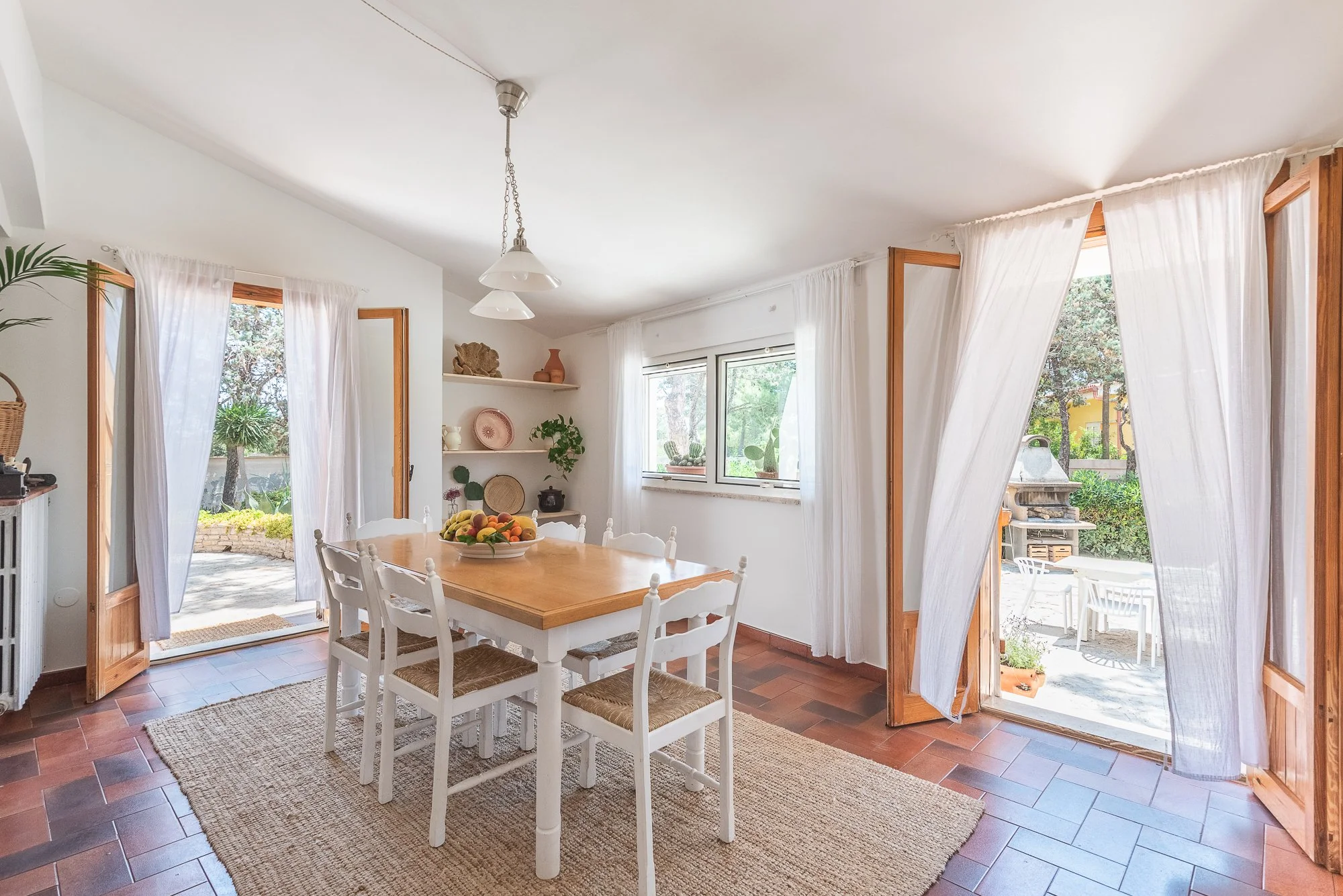 Ein helles Esszimmer mit weißen Wänden, einem Holztisch und weißen Stühlen, umgeben von großen Fenstern mit weißen Vorhängen, die zu einer Terrasse mit Garten führen.