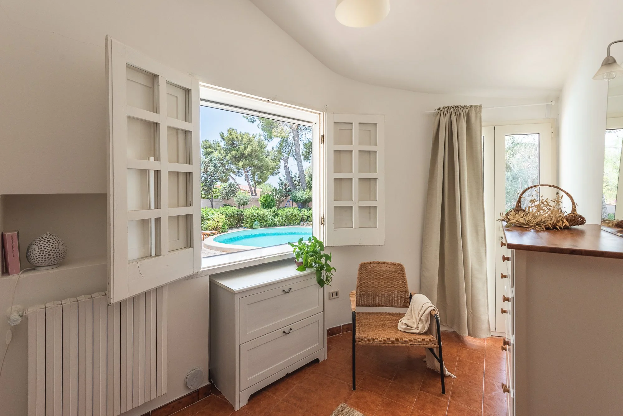 Ein helles Wohnzimmer mit offenem Fenster, Blick auf einen Pool im Garten, ein Stuhl mit Decke, weiße Möbel und ein kleiner Tisch, alles in warmen Farben.