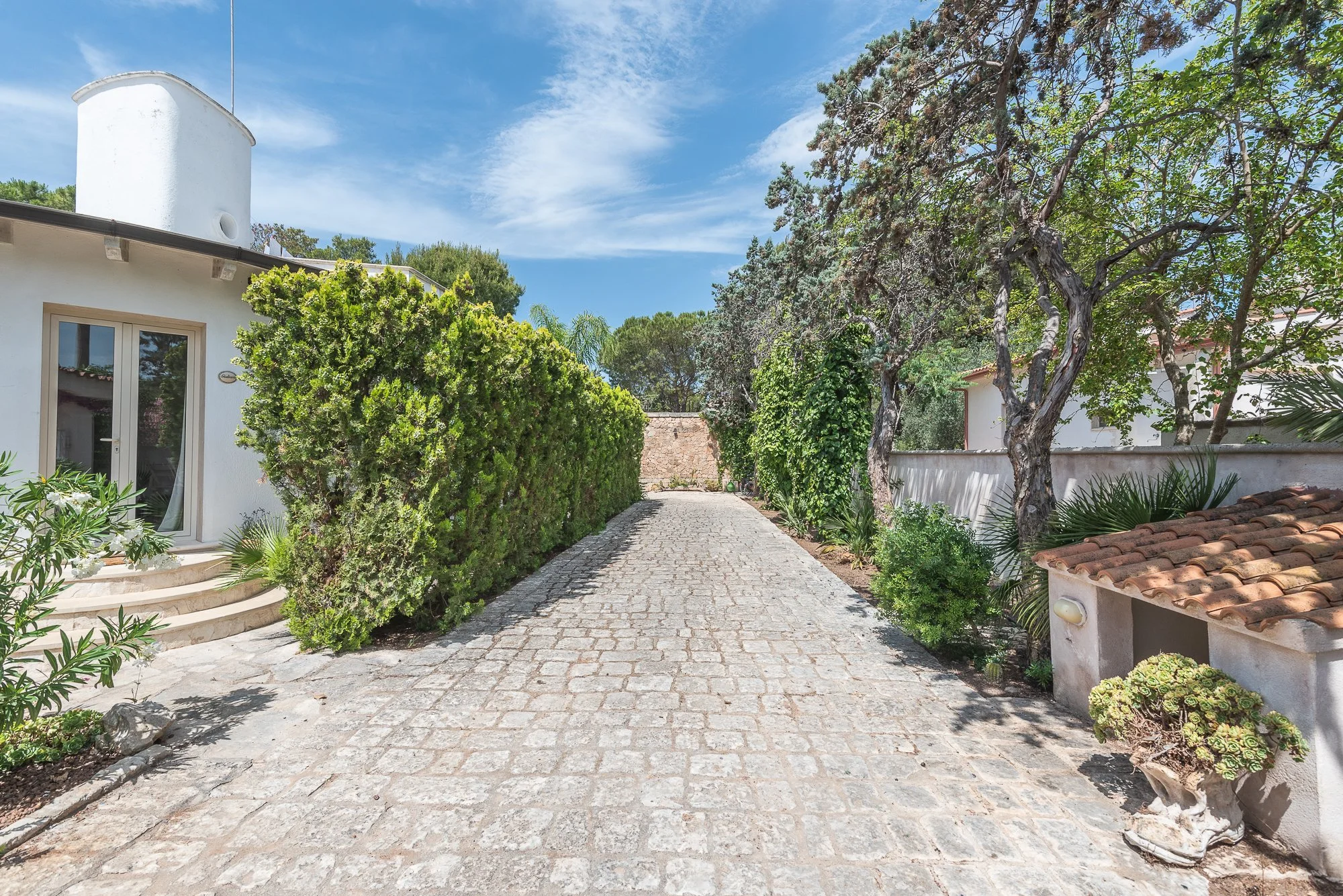 Ein Kiesweg führt durch einen grünen Garten mit Bäumen und Büsche auf beiden Seiten, umgeben von weißen Gebäuden mit Terrakottadächern und einer hohen weißen Mauer im Hintergrund. Der Himmel ist blau mit wenigen Wolken.