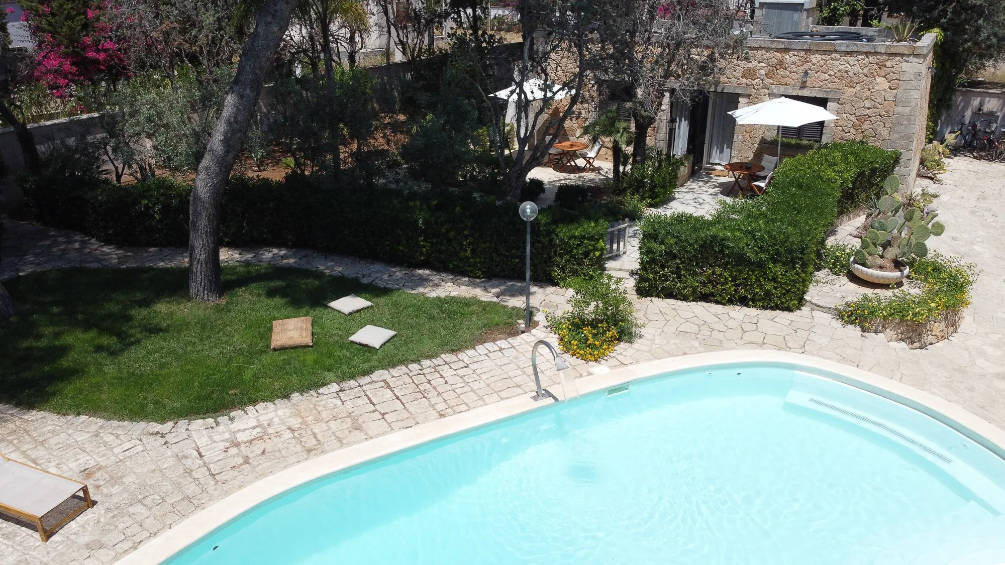 Ein Swimmingpool mit klarem Wasser, umgeben von Steinplatten, auf denen ein Liegestuhl steht. Im Hintergrund befinden sich Bäume, Büsche, eine kleine Gartenlaube mit Sitzgruppe, Pflanzen und ein Gepäckstück. Es ist sonnig und es gibt Schatten durch d