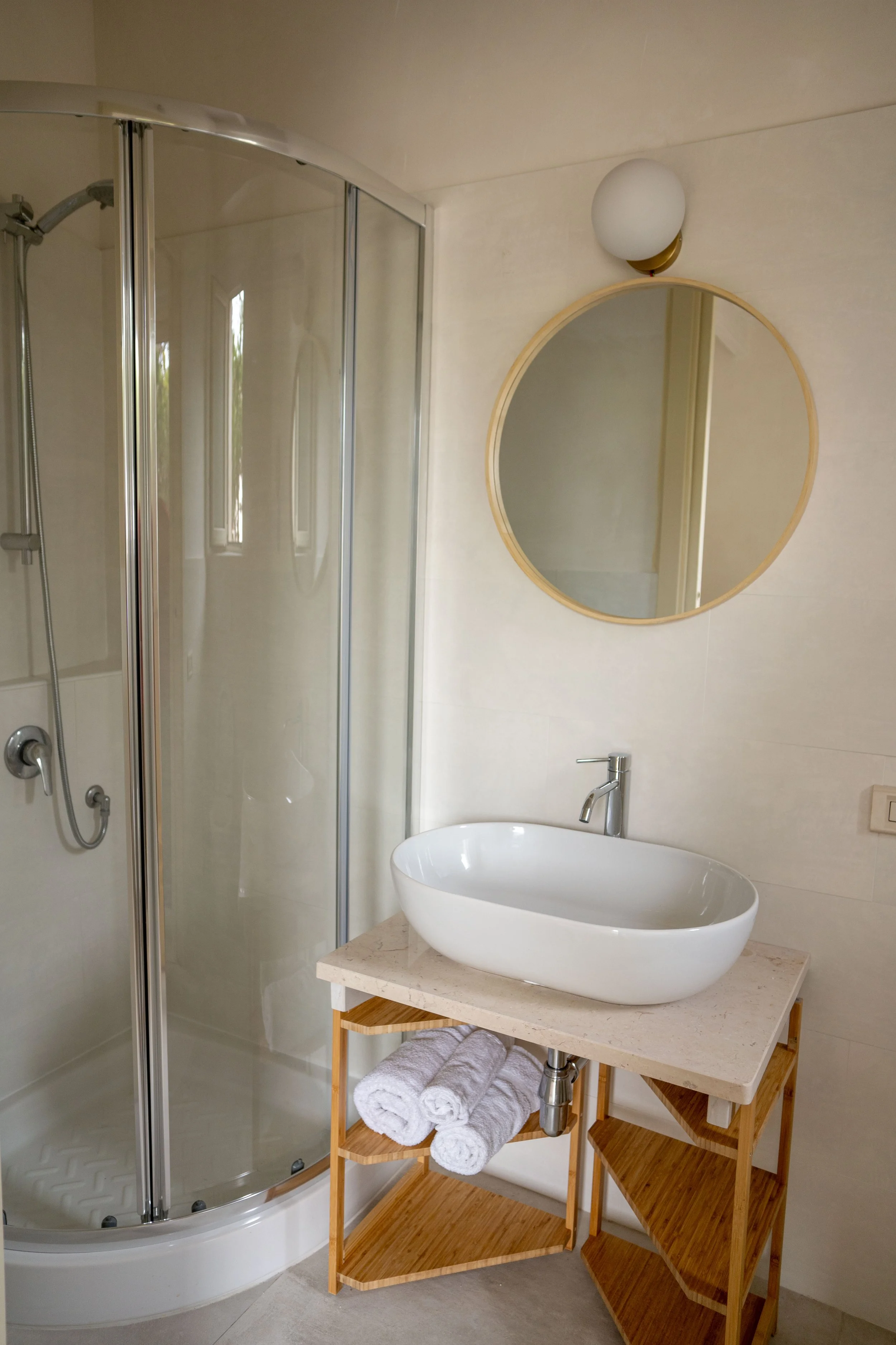 Ein Badezimmer mit einer Dusche, einem ovalen Waschbecken auf einem Holzregal und einem runden Spiegel an der Wand.