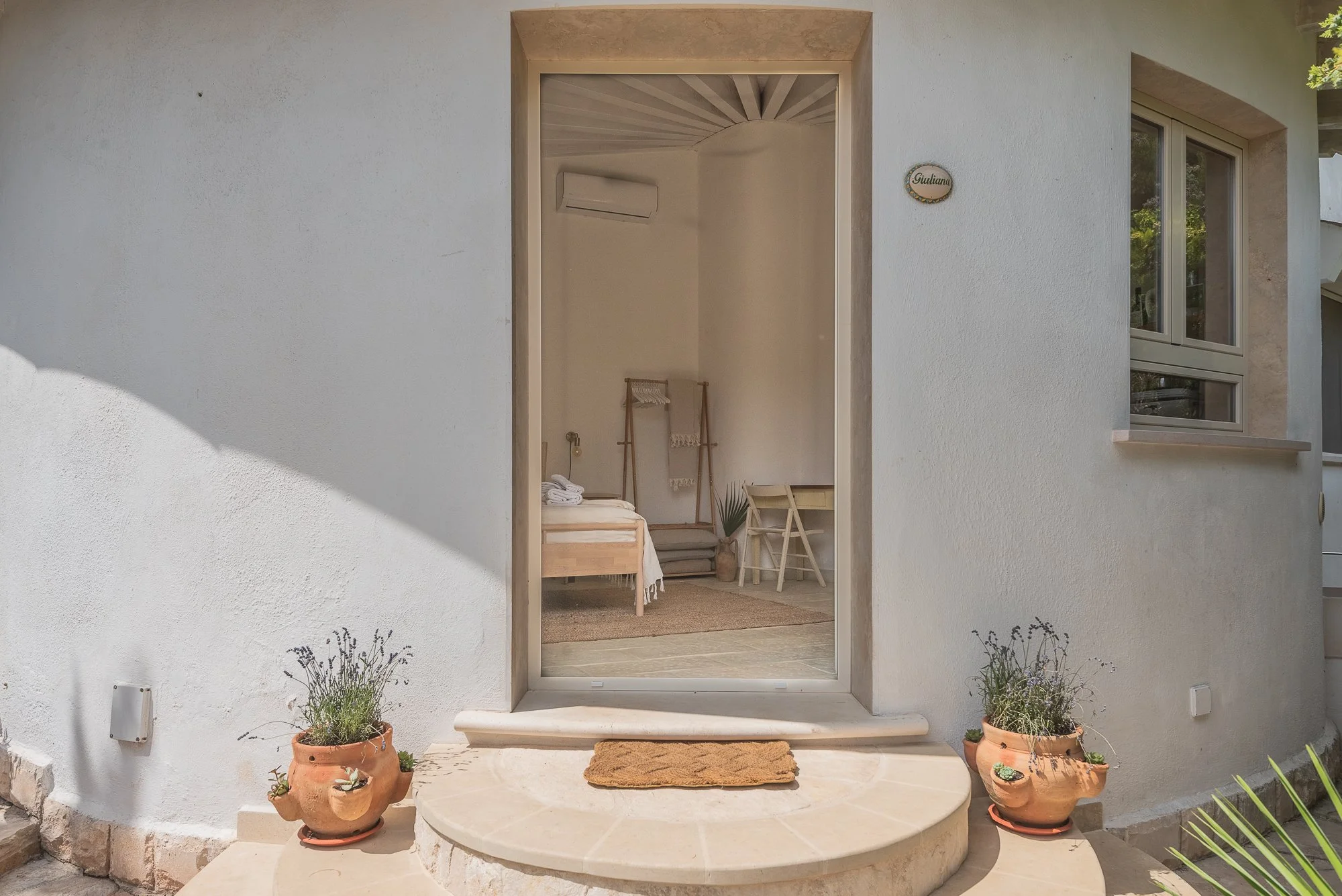 Blick durch eine offene Haustür ins Innere eines Schlafzimmers mit Bett, Stühlen und Dekorationen, außen davor Terrassentritt mit braunem Teppich, an beiden Seiten Terrakotta-Blumentöpfe mit Pflanzen, weiße Wand, ein Fenster und ein Namensschild mit 