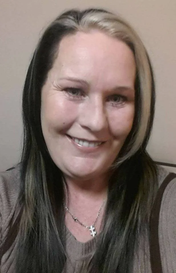 A woman with long black hair and blonde streaks, smiling, wearing a gray top and a cross necklace.