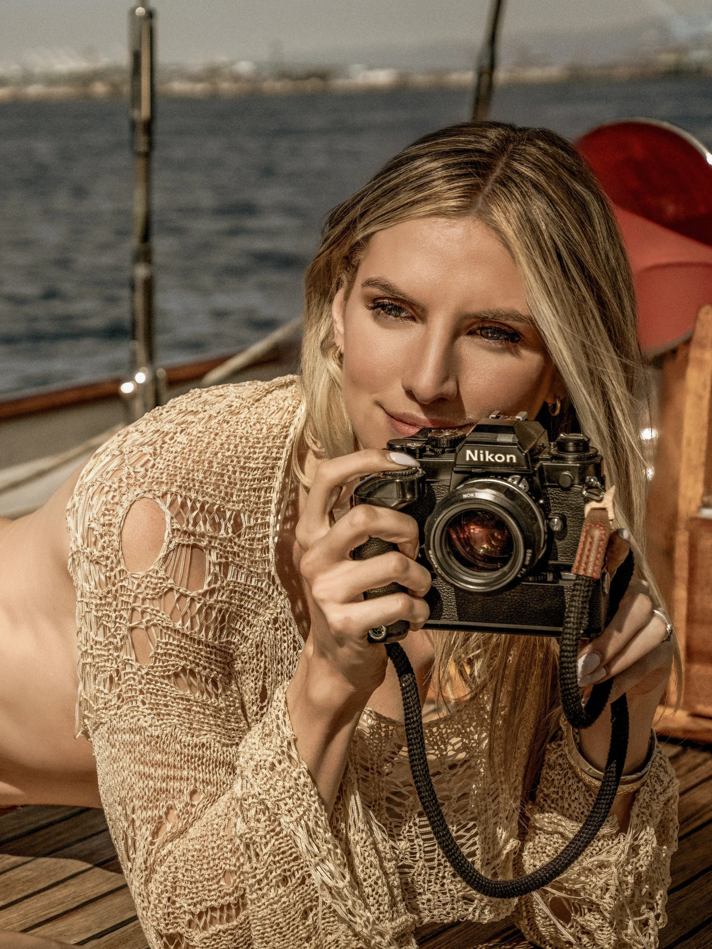 A woman with blonde hair taking a photograph with a Nikon camera on a boat with water and a distant shoreline in the background.