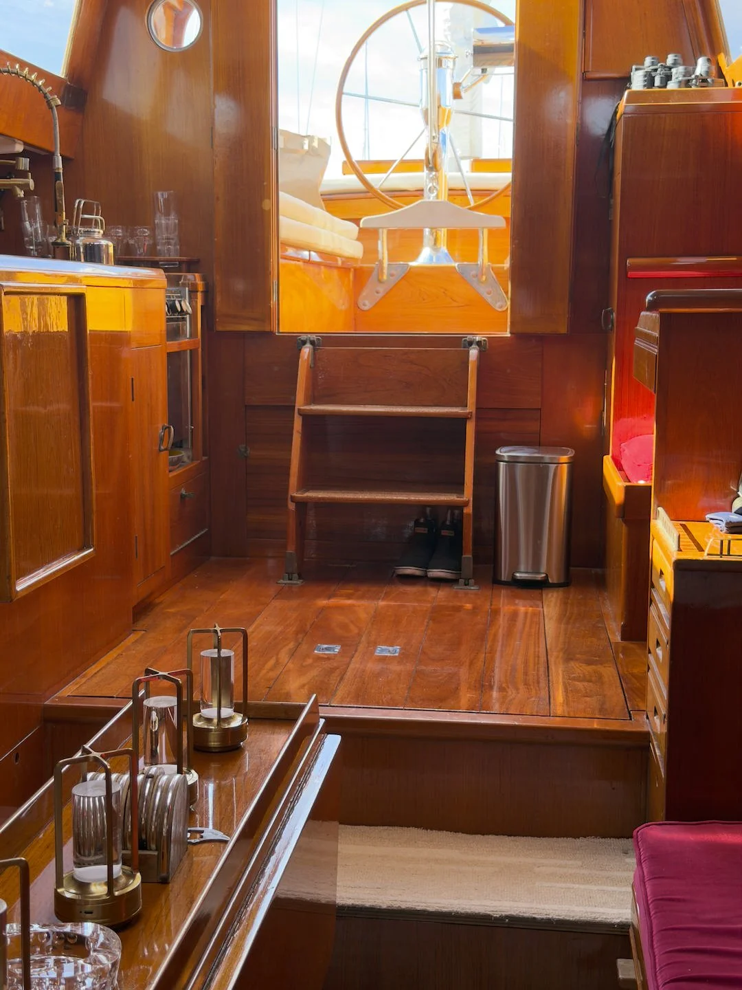 Interior of a boat's galley with wooden cabinets, a small step ladder, a window with a wheel outside, and various kitchen utensils and appliances.