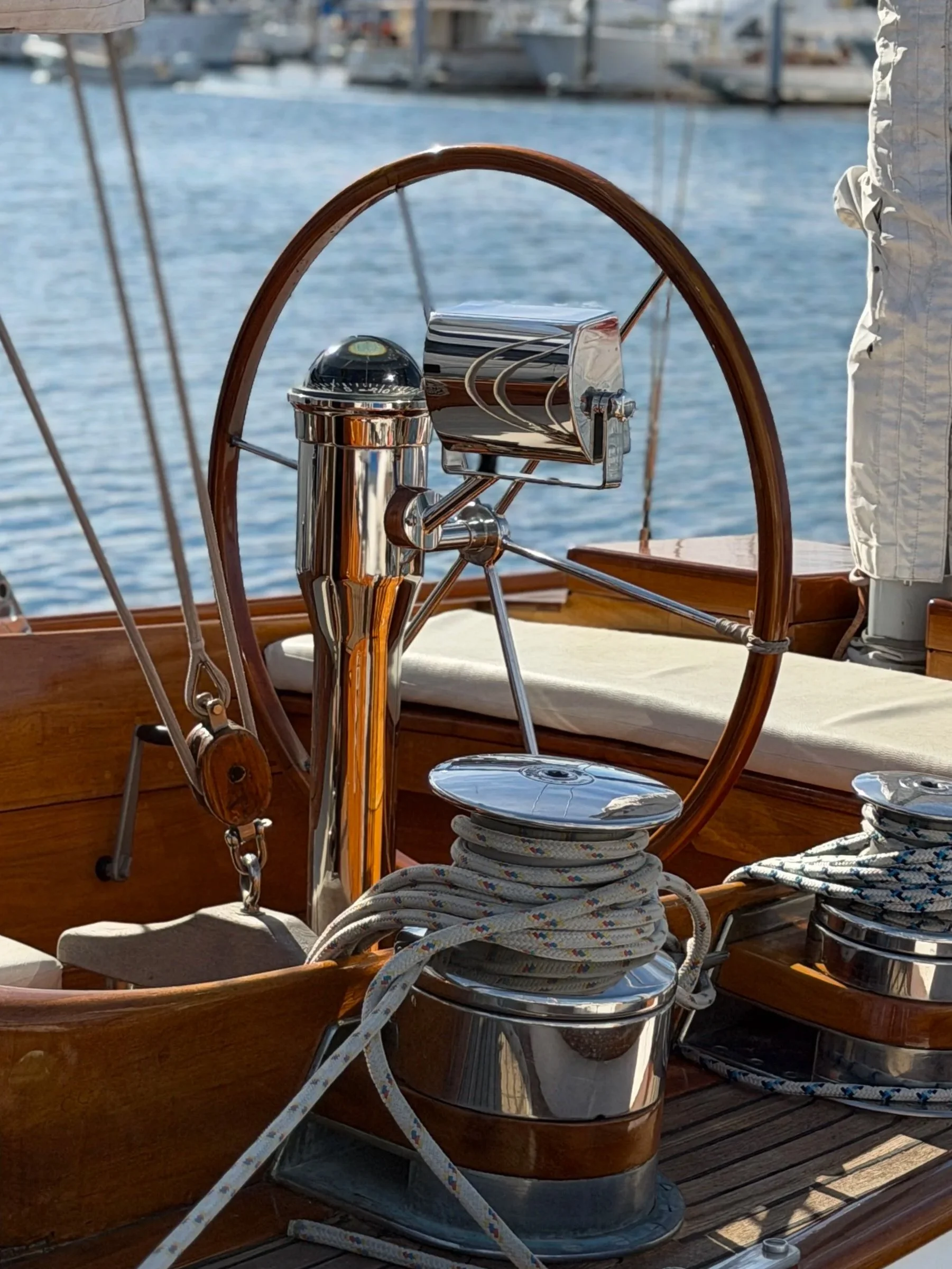 Close-up of a boat's steering wheel and winches with ropes, set against a water background.