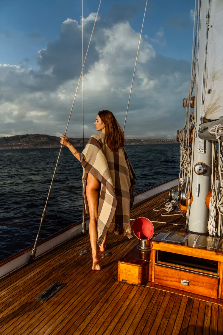 A woman standing barefoot on the deck of a sailboat, wrapped in a cashmere kaftan, holding a rope, with cloudy skies and water in the background.
