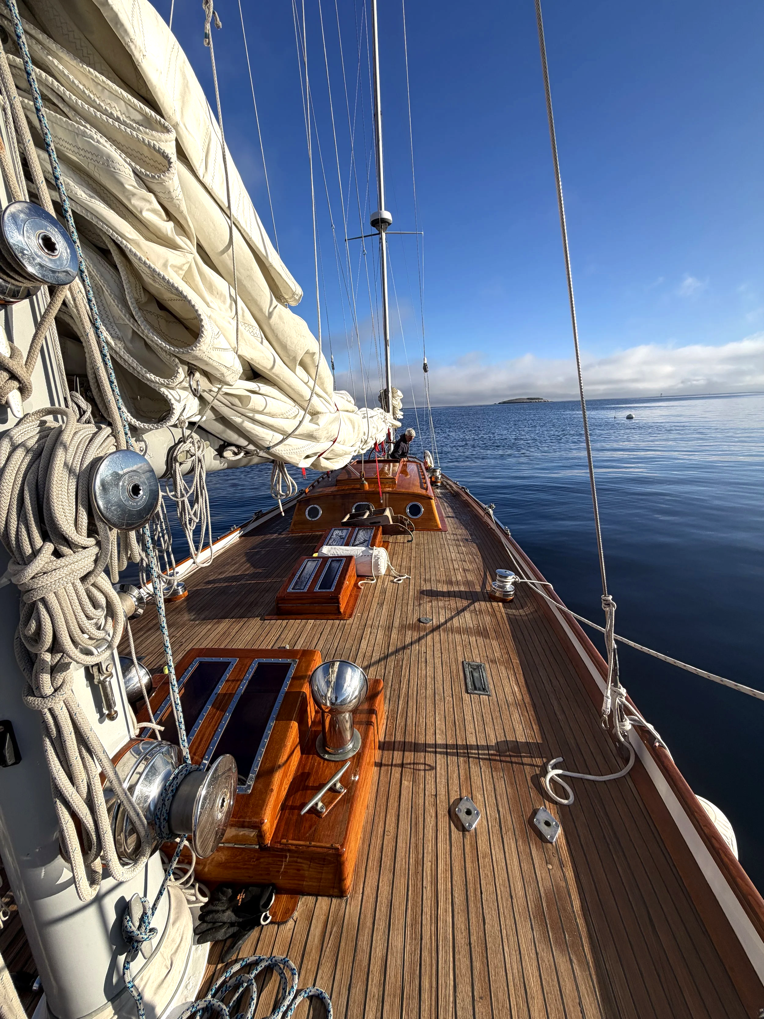 View of a wooden sailboat deck with coiled ropes, cleats, and their fittings, extending toward the blue sky and calm water with a distant small island on the horizon.