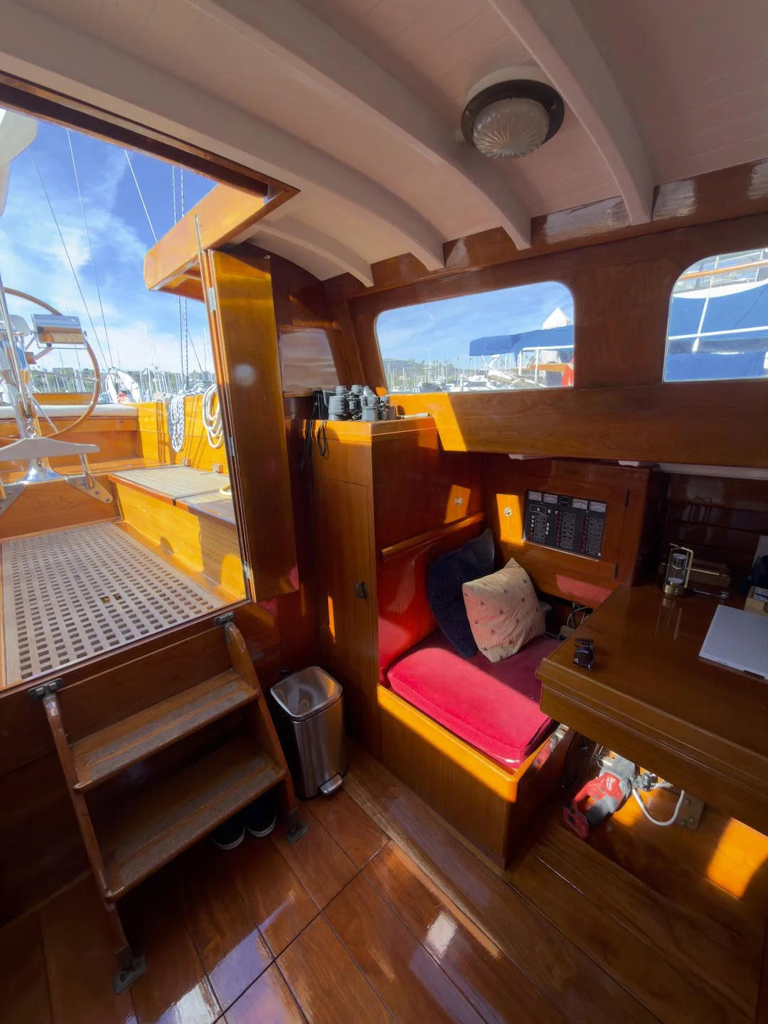 Interior of a boat with wooden paneling, a small seating area with cushions, a desk with electronic equipment, and a window showing external boats and blue sky.