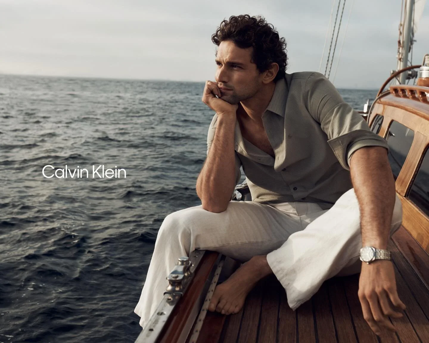 Man sitting on a boat, looking out at the ocean, wearing a light-colored Calvin Klein shirt and pants, with a watch on his wrist, during daytime.