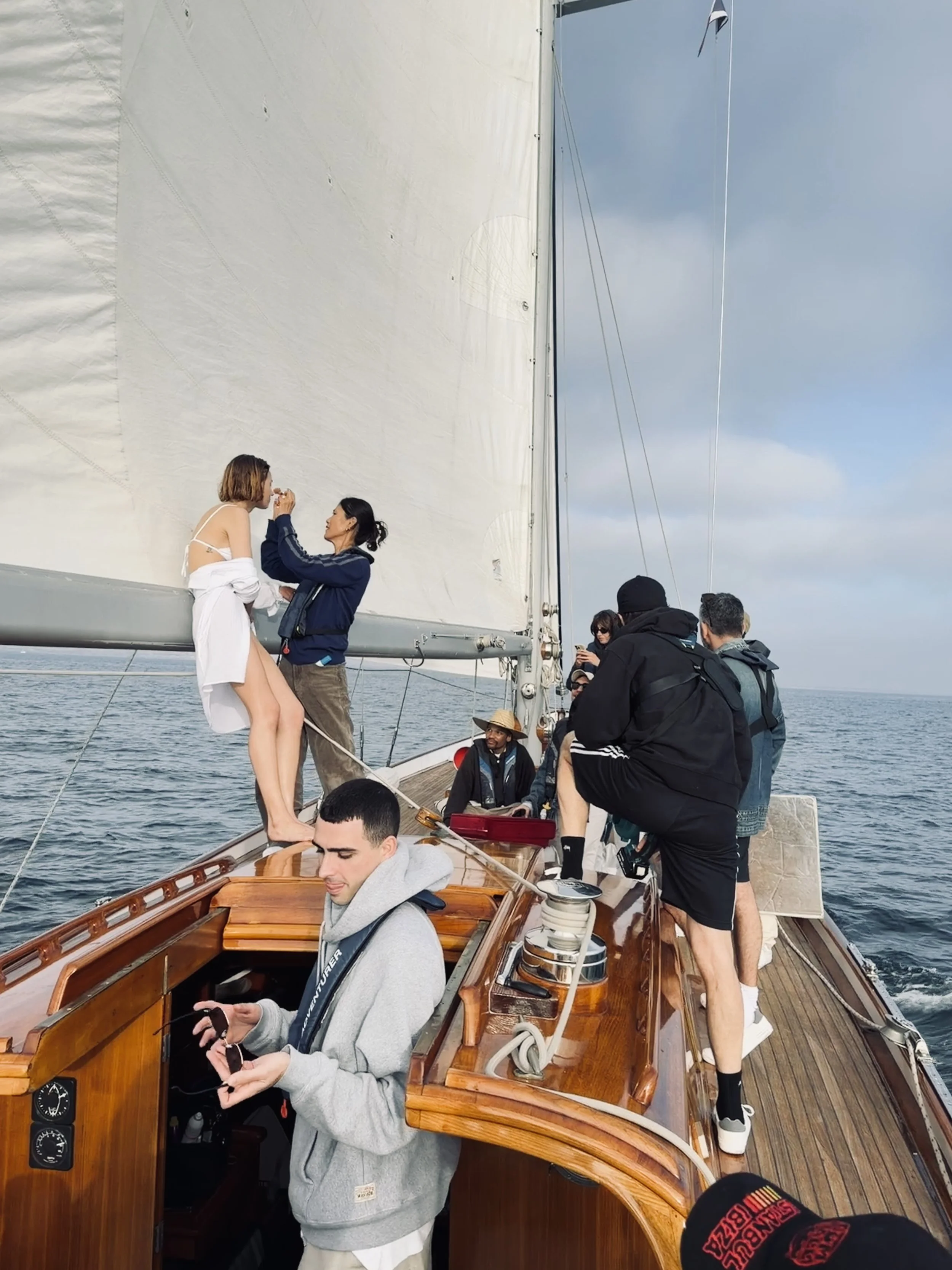 Group of people sailing on a boat, some are working with ropes and others are relaxing, with a woman sitting on the edge of the boat and a woman standing nearby taking a photo, under an overcast sky.