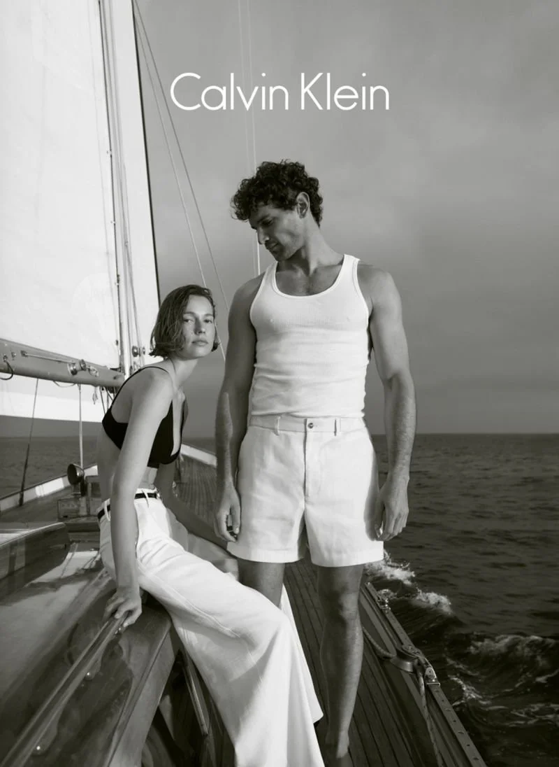 Black and white photo of a man and woman on a boat with ocean in background, both dressed in casual summer clothes, with the text 'Calvin Klein' at the top.