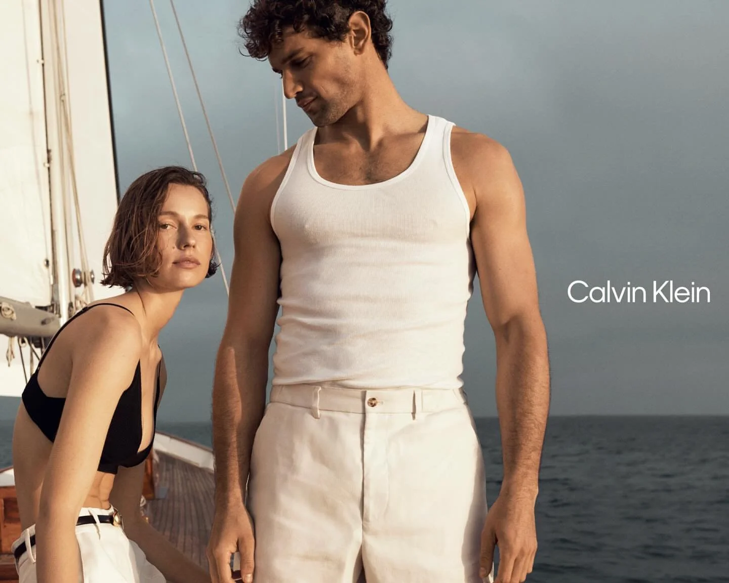 A man and woman standing on a boat deck by the ocean. The woman has short, wavy hair and wears a black crop top and white pants. The man has curly hair and wears a white tank top and off-white pants. The sky is overcast. The text "Calvin Klein" appea