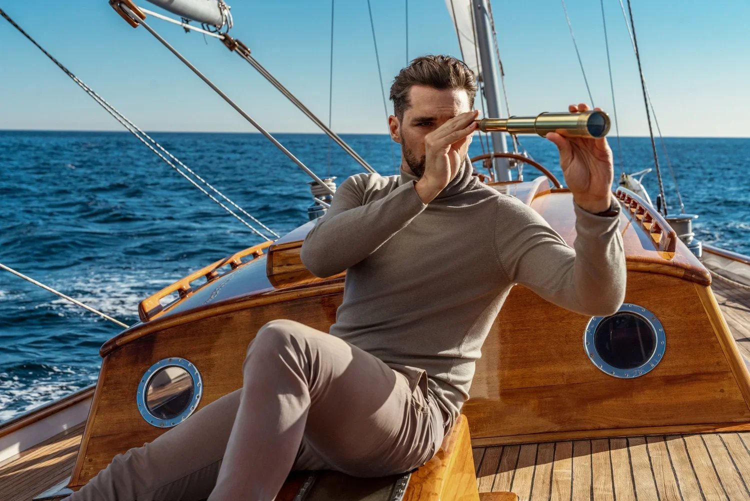 Man on a sailboat looking through a spyglass with the ocean in the background.