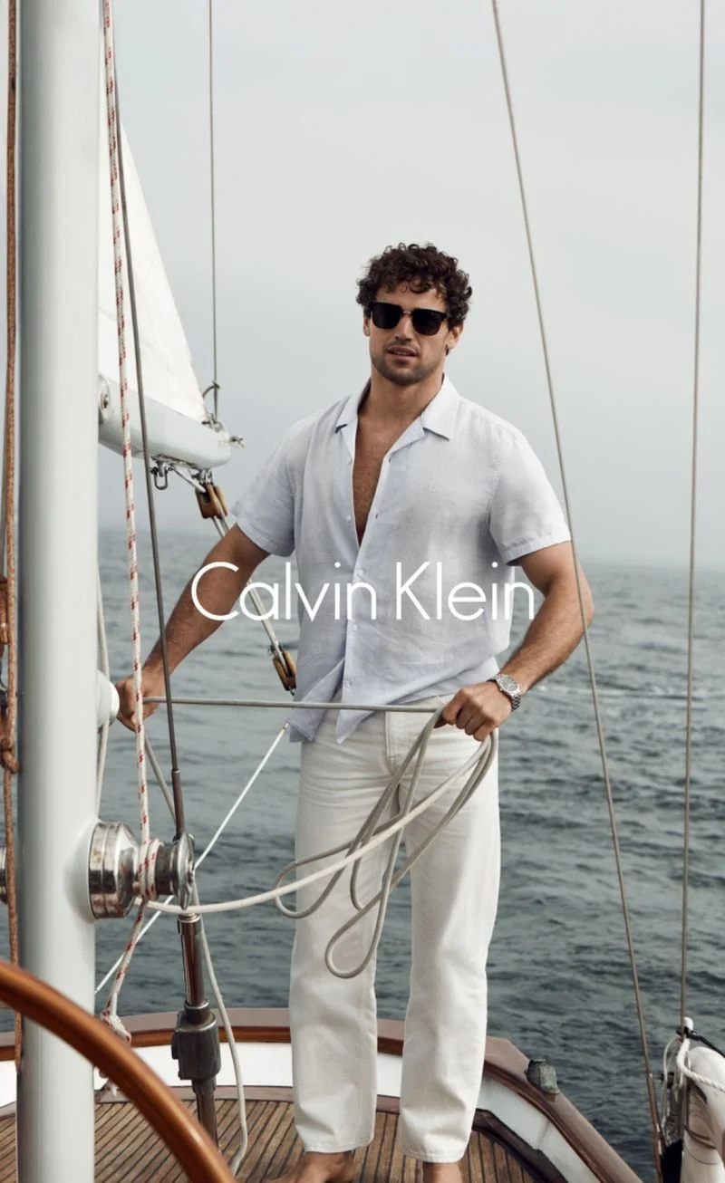 A man with curly brown hair, wearing sunglasses, a white short-sleeve shirt, and white pants, stands on a sailboat holding a rope with the ocean in the background. The text 'Calvin Klein' is overlaid on the image.